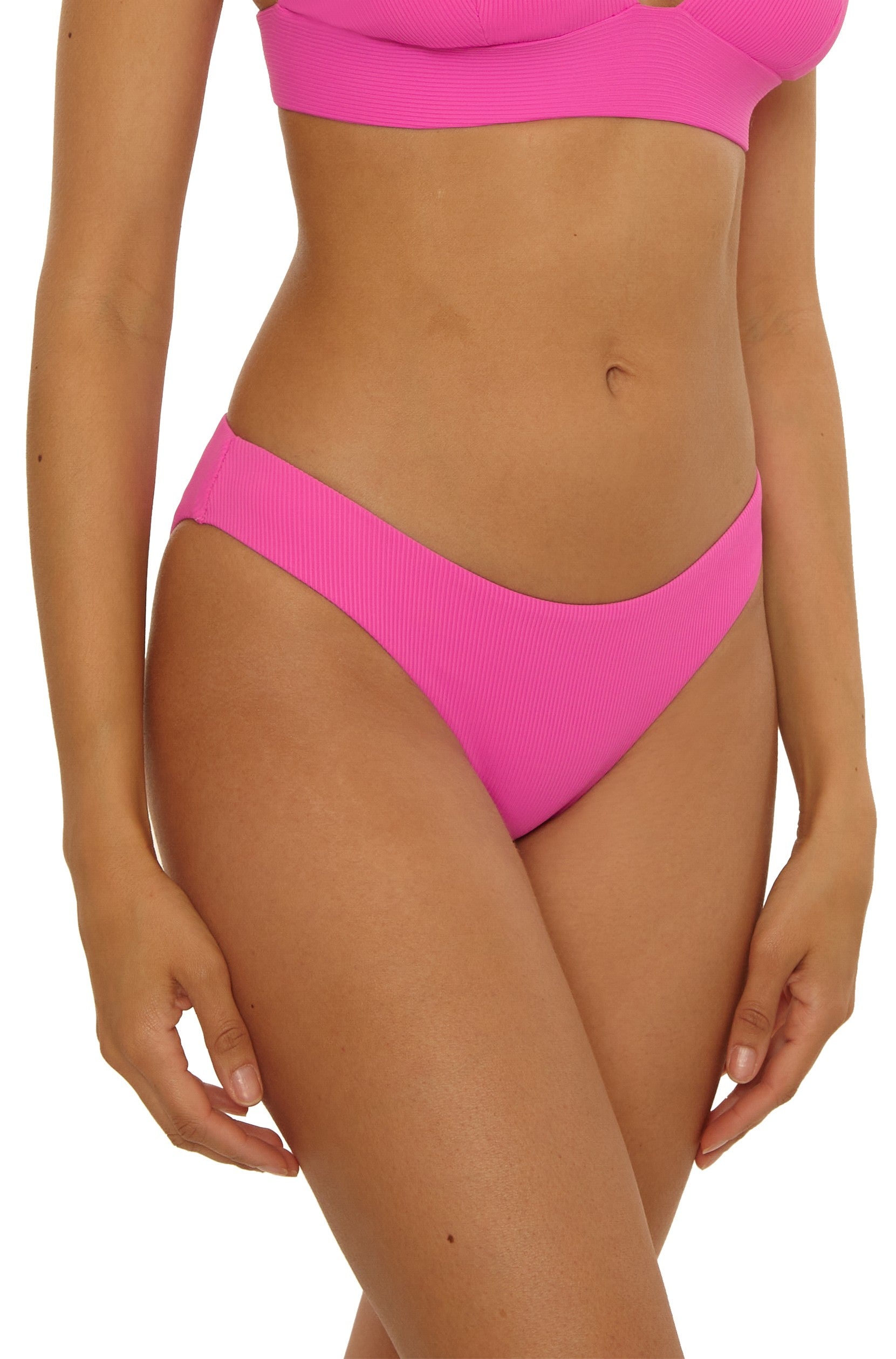 A woman models the Becca Swim Modern Edge Hipster bikini in pink, featuring a fine rib texture and hipster bottom, shown from mid-chest to upper thighs with her hand by her side against a plain background.
