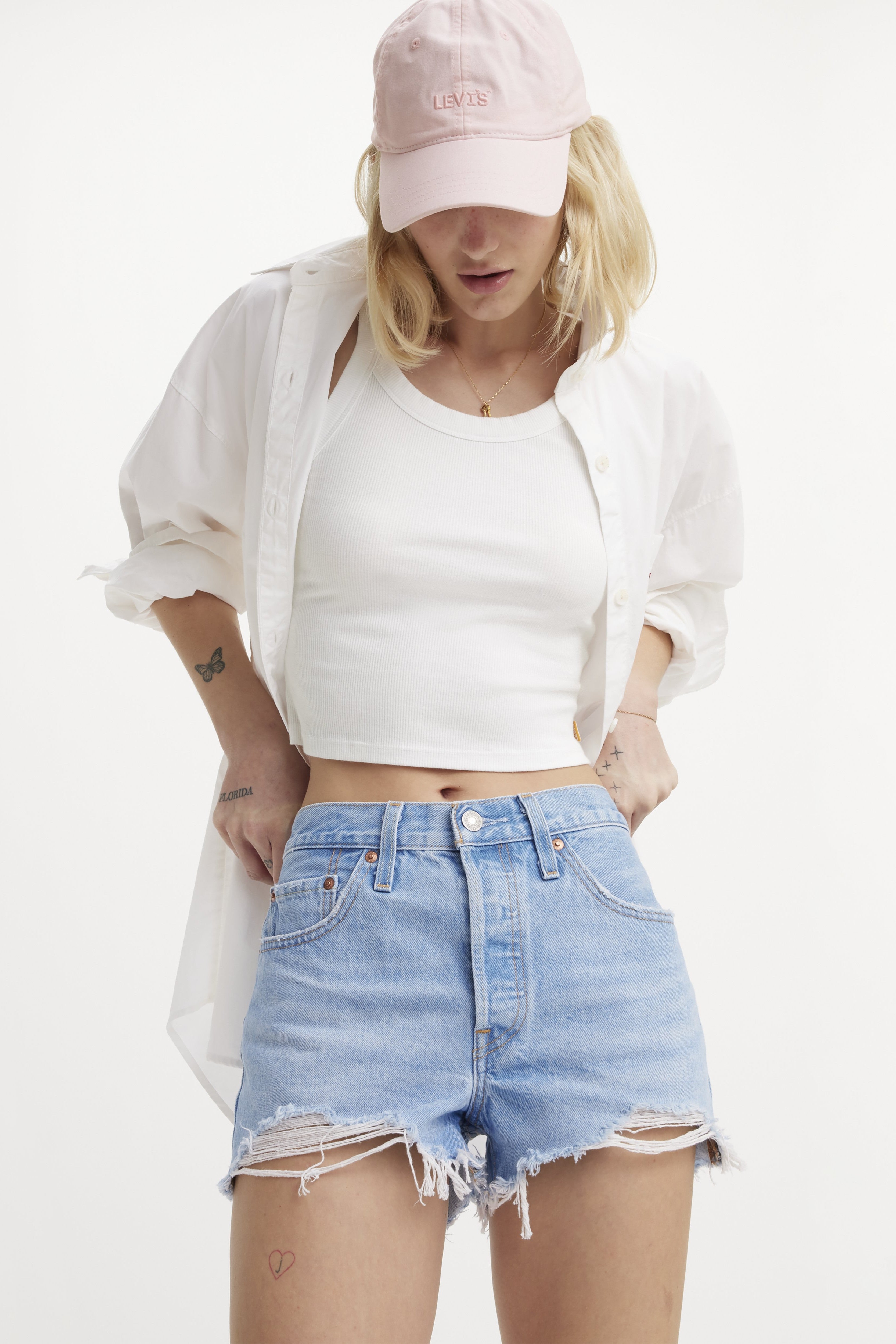 A person stands against a plain white background wearing a light pink cap, white crop tank, unbuttoned white shirt, and Levi's 501 High Rise Cut Off Denim Short in Luxor Heat.