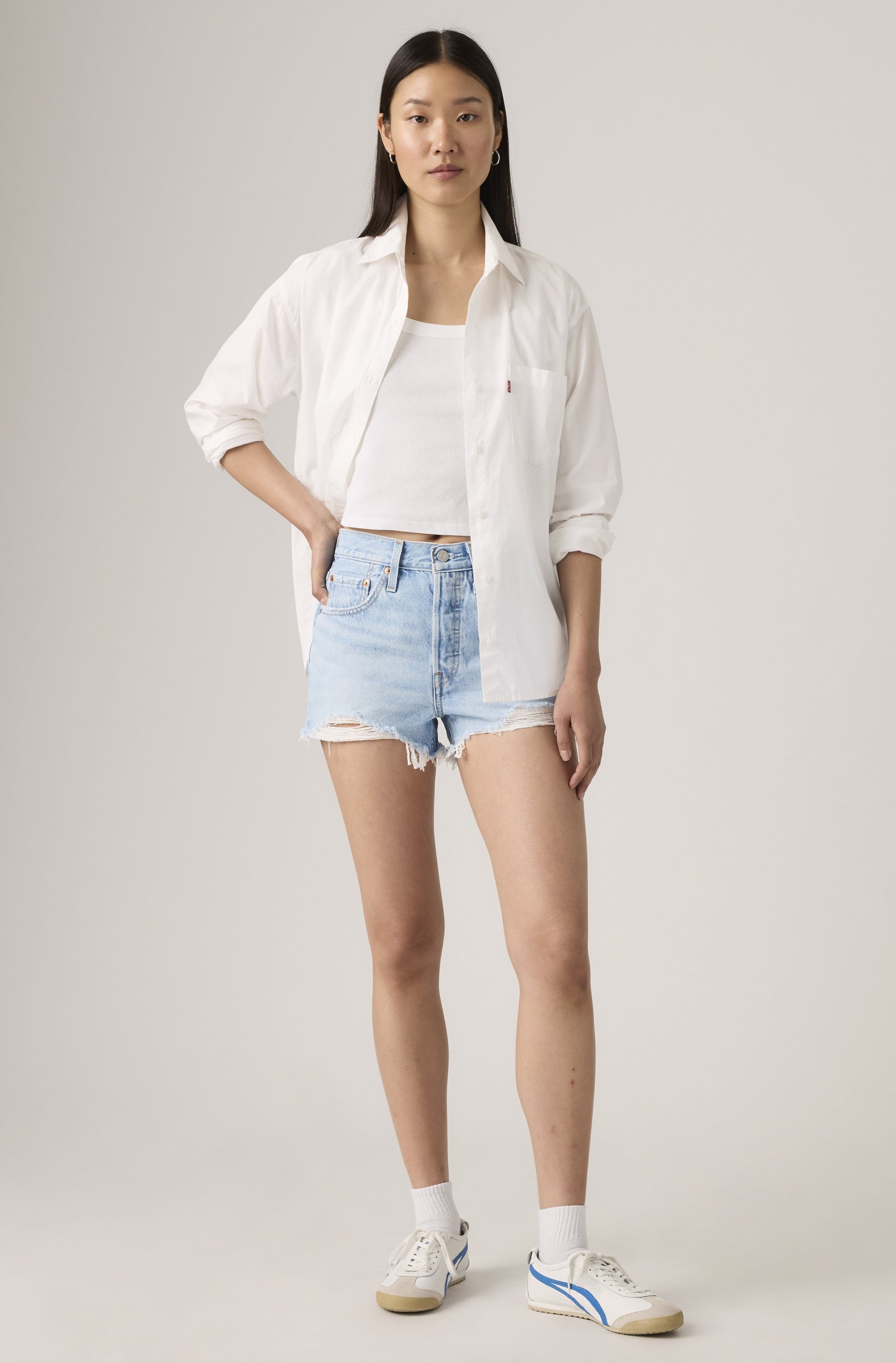 A woman with long, straight hair and a neutral expression wears Levi’s 501 High Rise Cut Off Denim Short in Luxor Heat, a white button-up shirt over a white tank top, white socks, and white sneakers against a plain background.