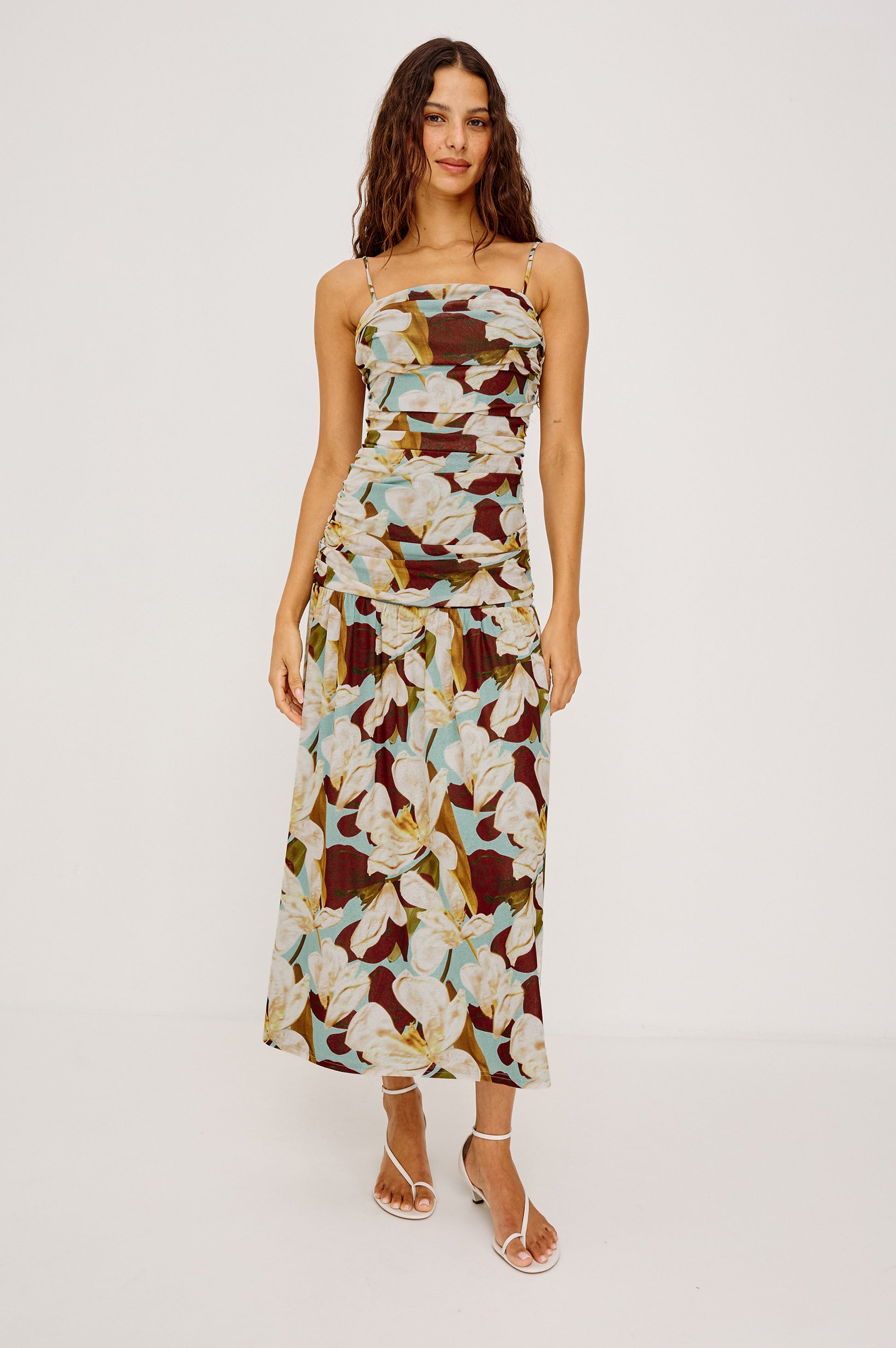 A woman wears the Rails Dolly Midi Dress, a timeless floral-print midi in white, yellow, and green on a dark base, paired with white strappy sandals against a plain background.
