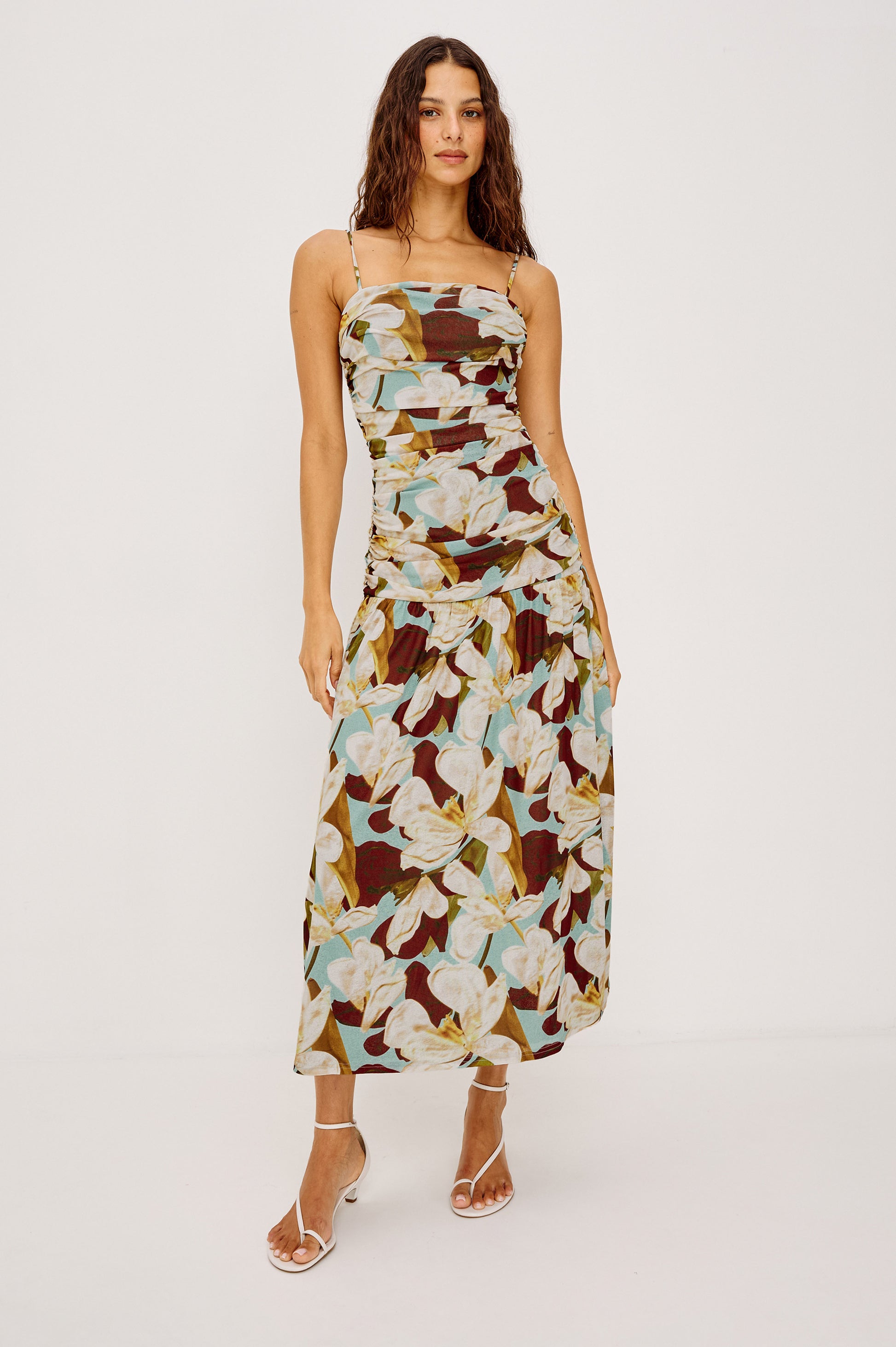 A woman poses against a white background in the Rails Dolly Midi Dress in Brown Multi, featuring a sleeveless, ruched silhouette with bold white floral print and styled with white strappy sandals.