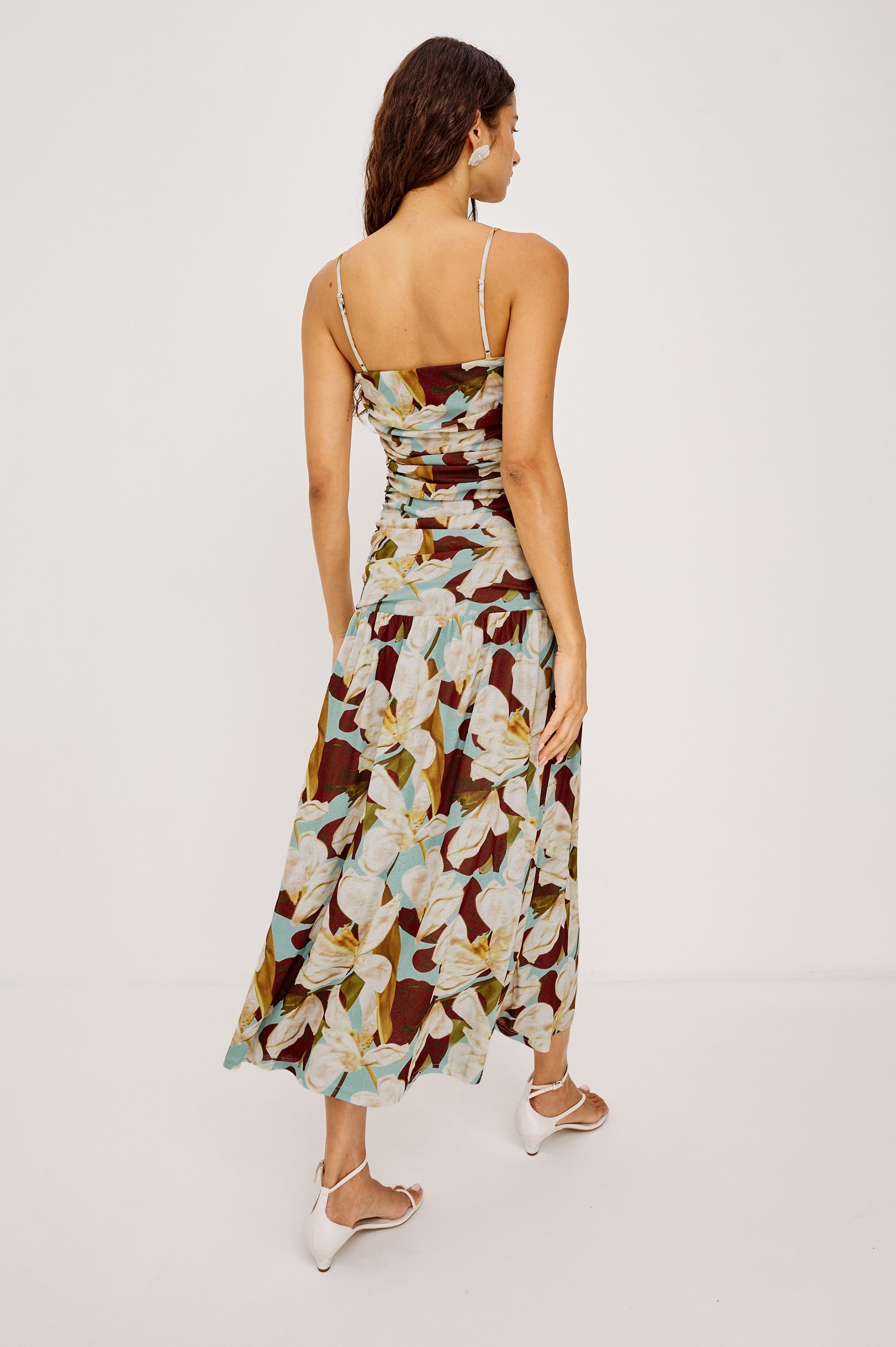 A woman faces away, wearing the Rails Dolly Midi Dress—a fitted, sleeveless brown and white floral midi—paired with white heeled sandals against a plain light backdrop.