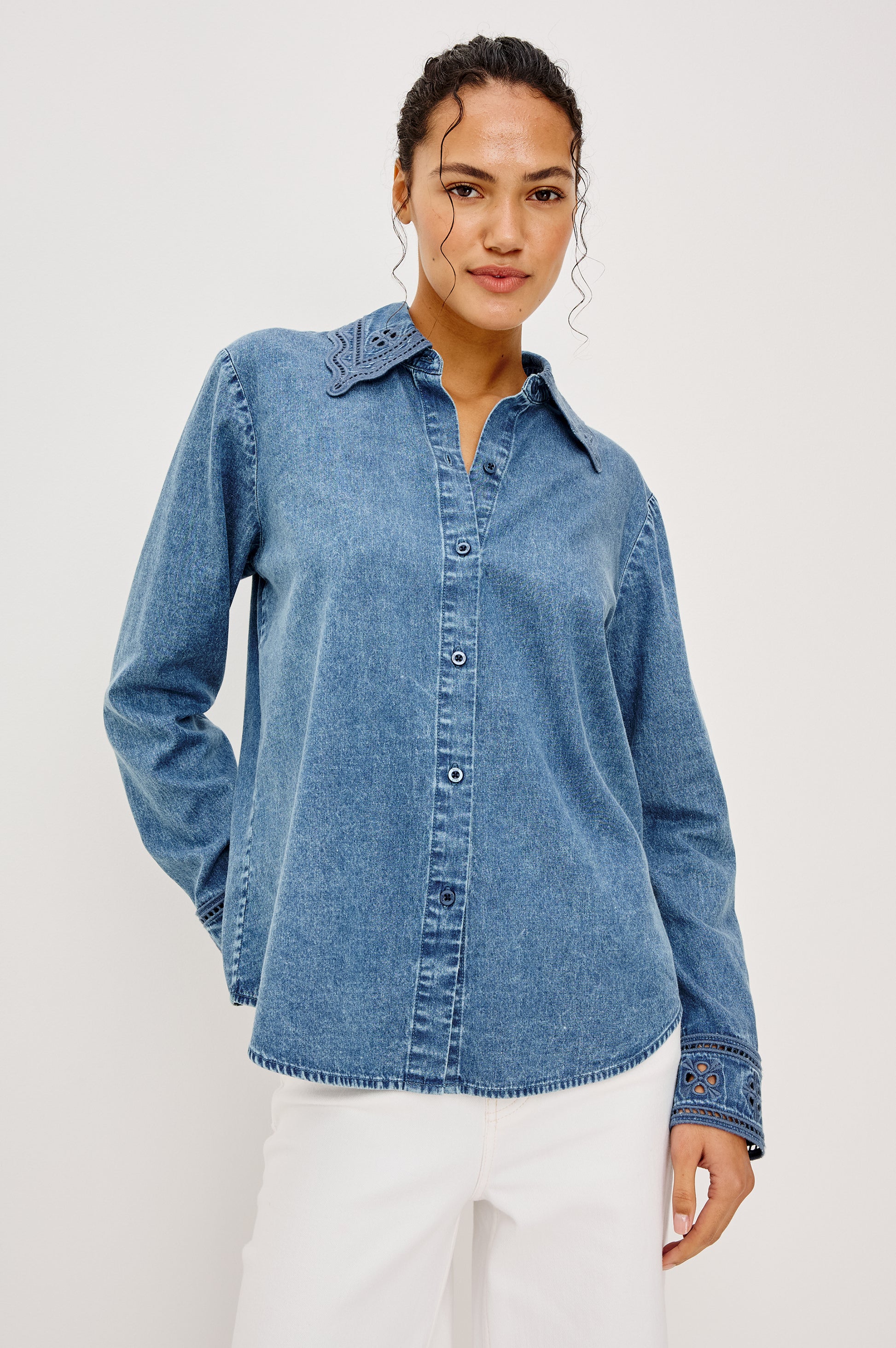 A woman smiles slightly, one hand in her pocket, wearing the Bethany Denim Long Sleeve Top by Rails—featuring medium-wash denim with embroidered collar and cuffs—paired with white pants, against a plain background.