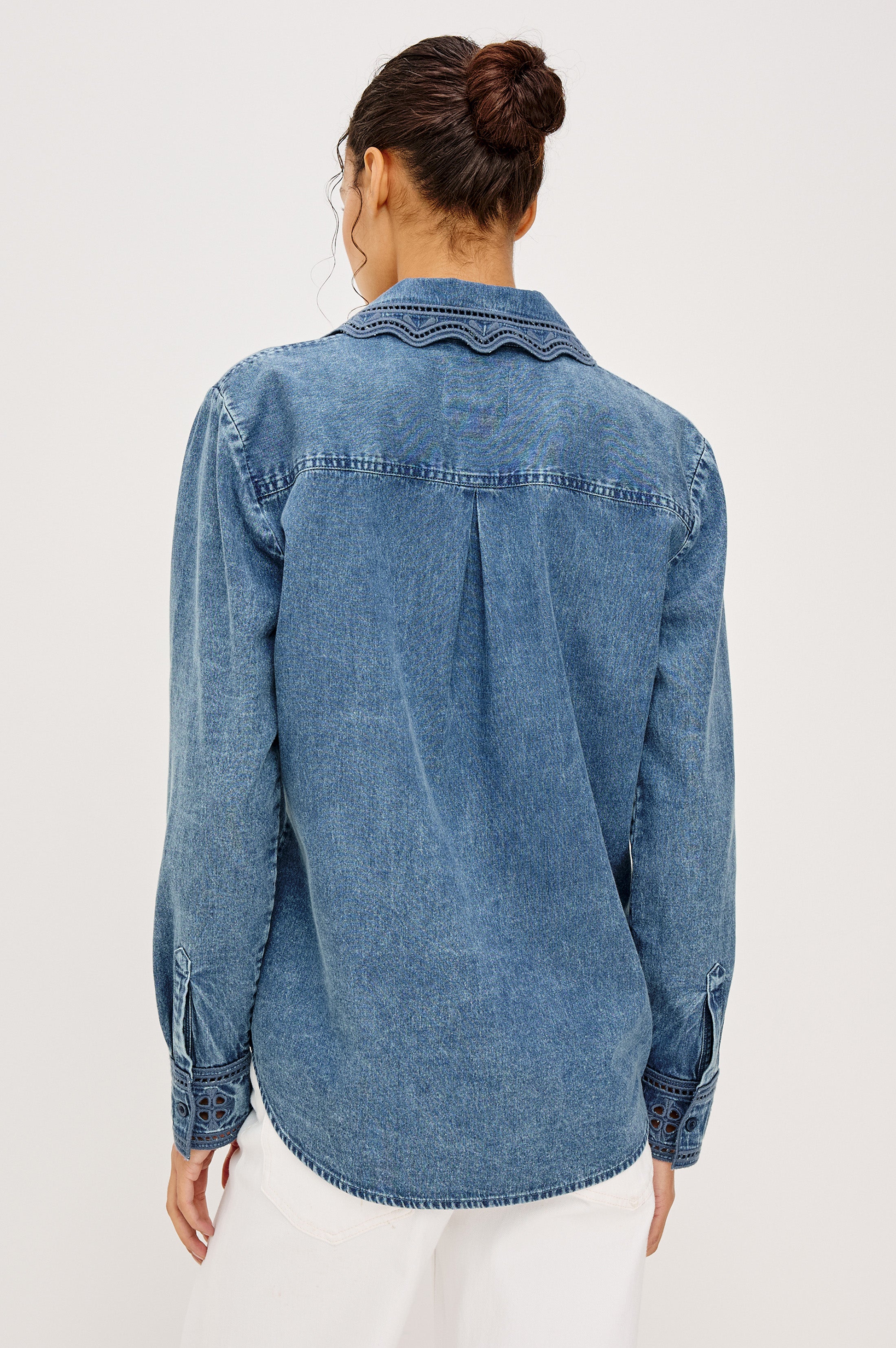 A person with dark hair in a bun is shown from the back, wearing the Rails Bethany Denim Long Sleeve Top and white pants, standing against a plain light background.