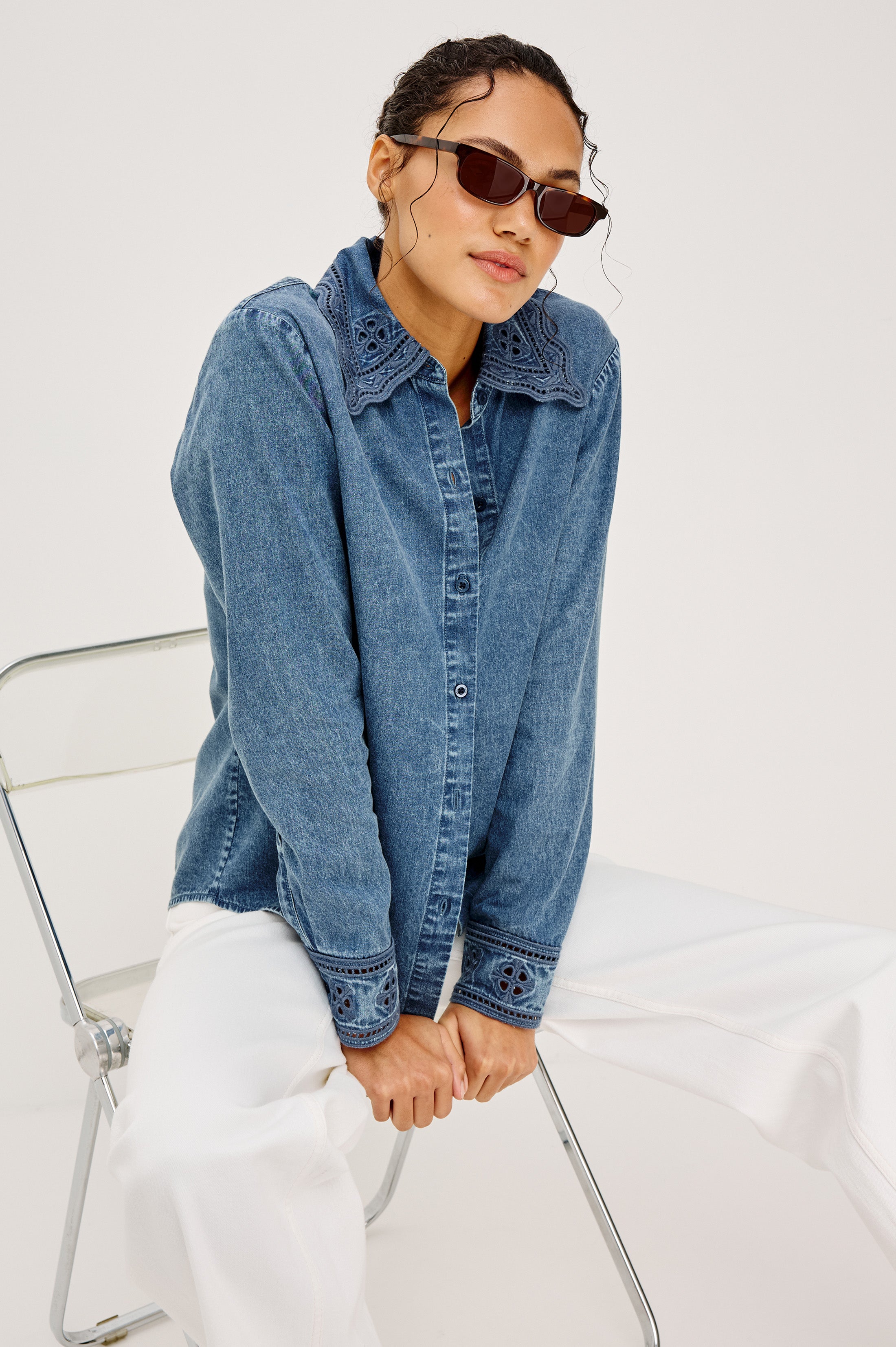 A person in the Rails Bethany Denim Long Sleeve Top and white pants sits casually on a transparent chair against a plain light background, accessorized with sunglasses.