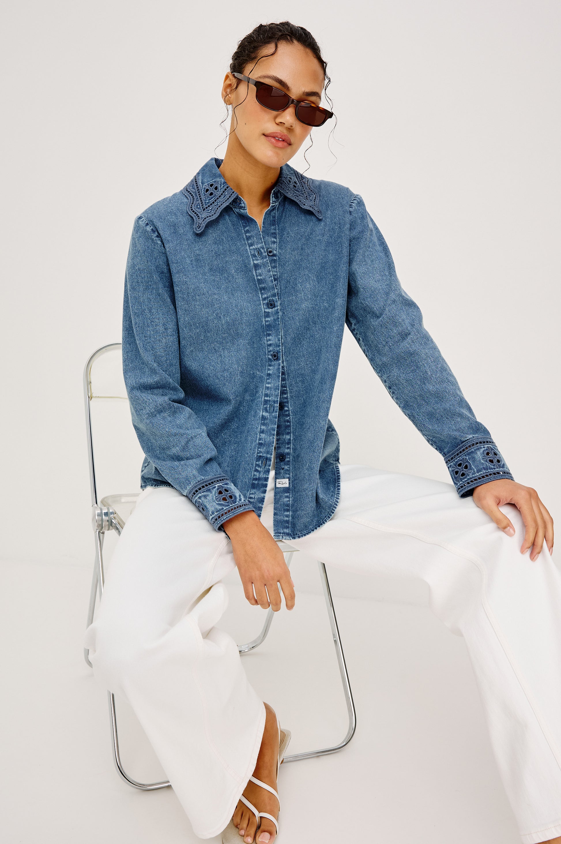 A woman wearing sunglasses and the Rails Bethany Denim Long Sleeve Top with embroidered details, paired with white wide-leg pants, sits confidently on a metal chair against a plain white background, one hand resting on her knee.