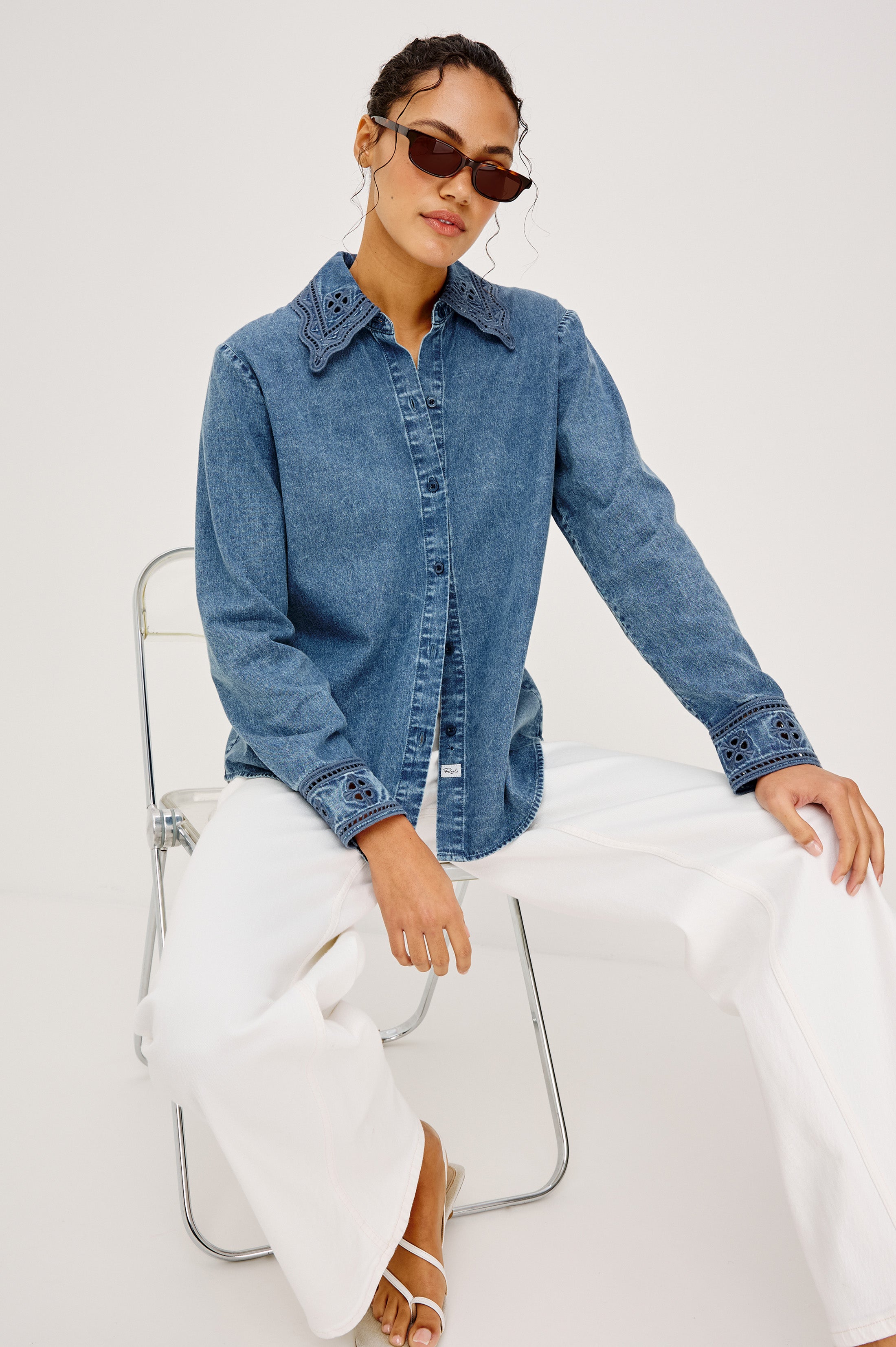 A woman wearing sunglasses and the Rails Bethany Denim Long Sleeve Top with embroidered details, paired with white wide-leg pants, sits confidently on a metal chair against a plain white background, one hand resting on her knee.