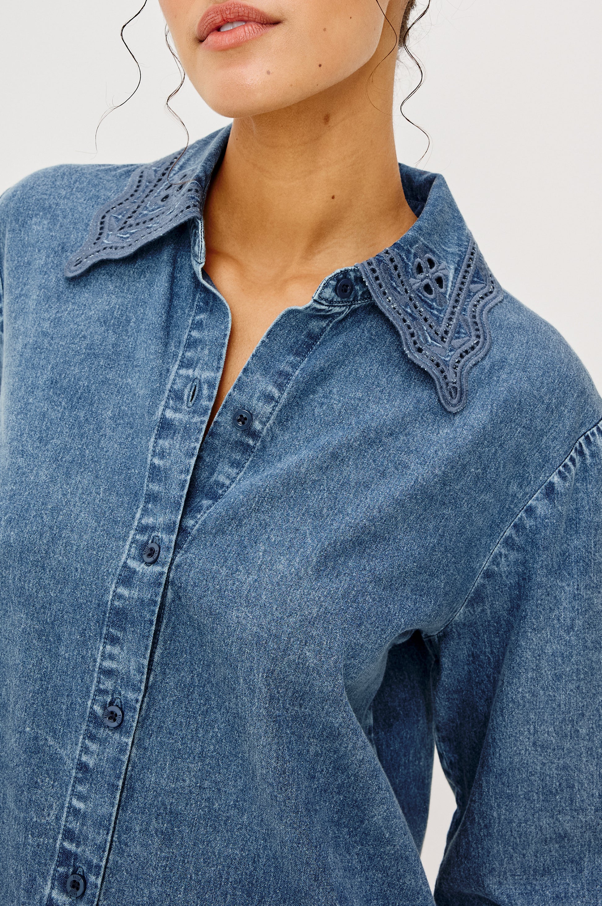 A woman wears the Rails Bethany Denim Long Sleeve Top, featuring a medium wash, button-down front, and an embroidered scalloped collar. Her face is partially visible and her hair falls in loose curls.