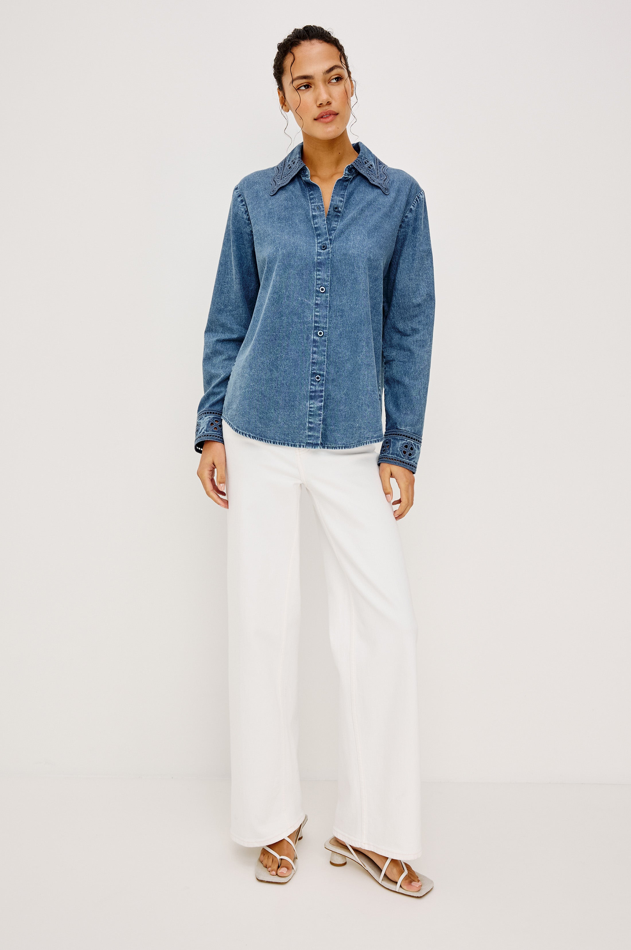 A woman stands against a plain light background, wearing the Rails Bethany Denim Long Sleeve Top, wide-leg white pants, and white sandals. Her hair is tied back and her hands rest calmly by her sides.