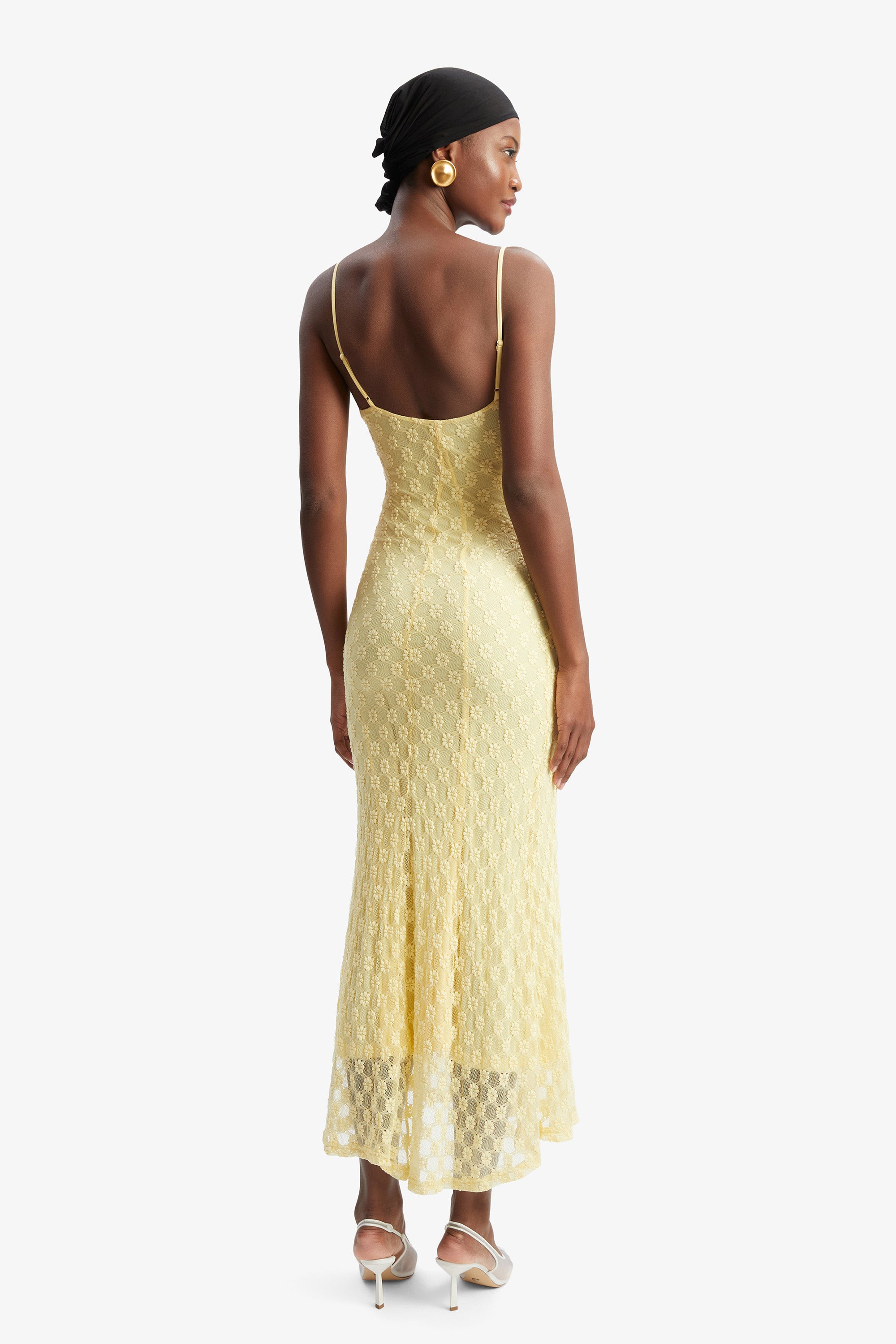 A woman in the Bardot Adoni Mesh Midi Dress and white heels stands facing away, wearing a black headscarf and gold hoop earrings, with her hands at her sides against a plain white background.
