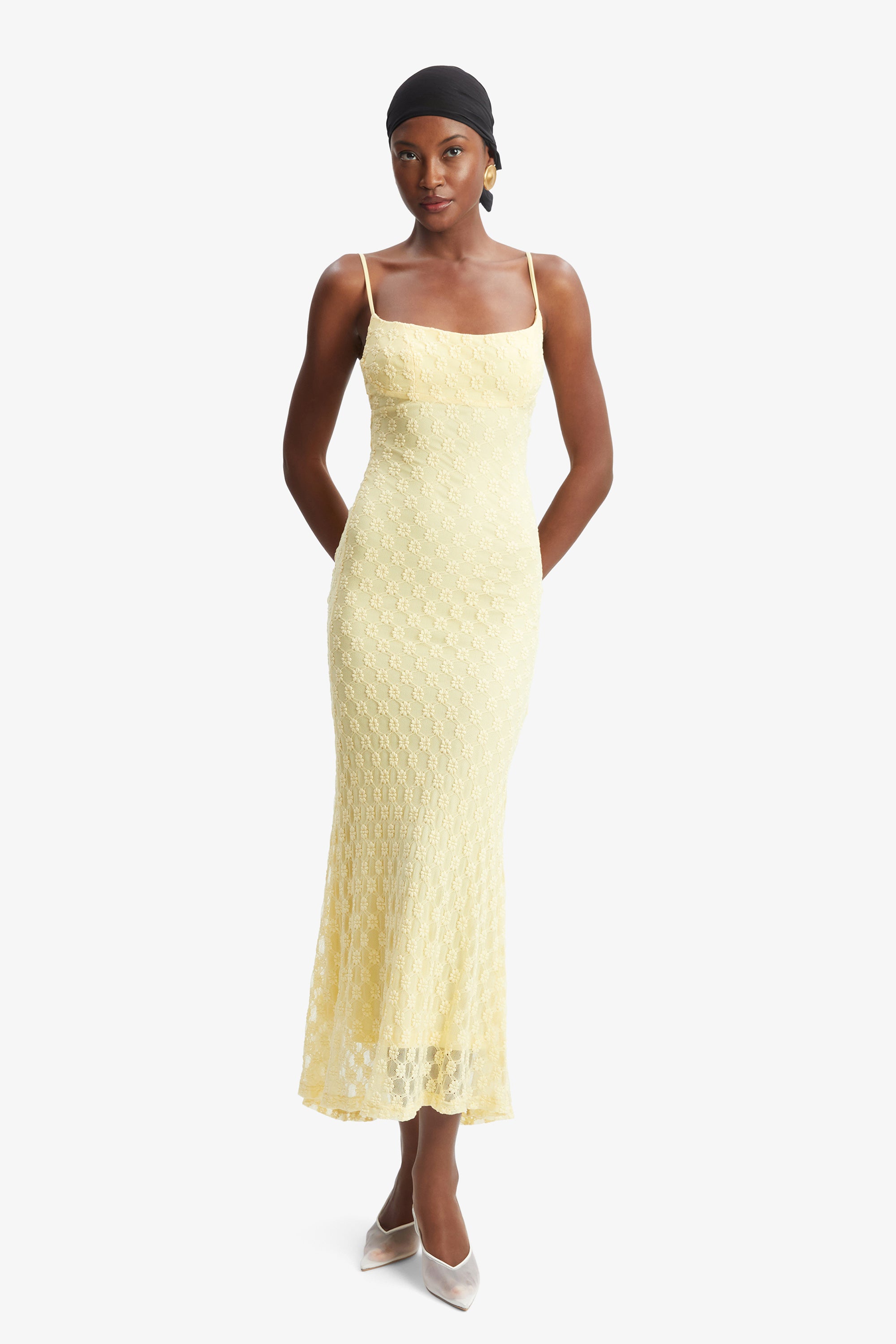 A woman stands against a plain white background wearing the Bardot Adoni Mesh Midi Dress in pale yellow, styled with spaghetti straps, white heels, gold hoop earrings, and a black headscarf.