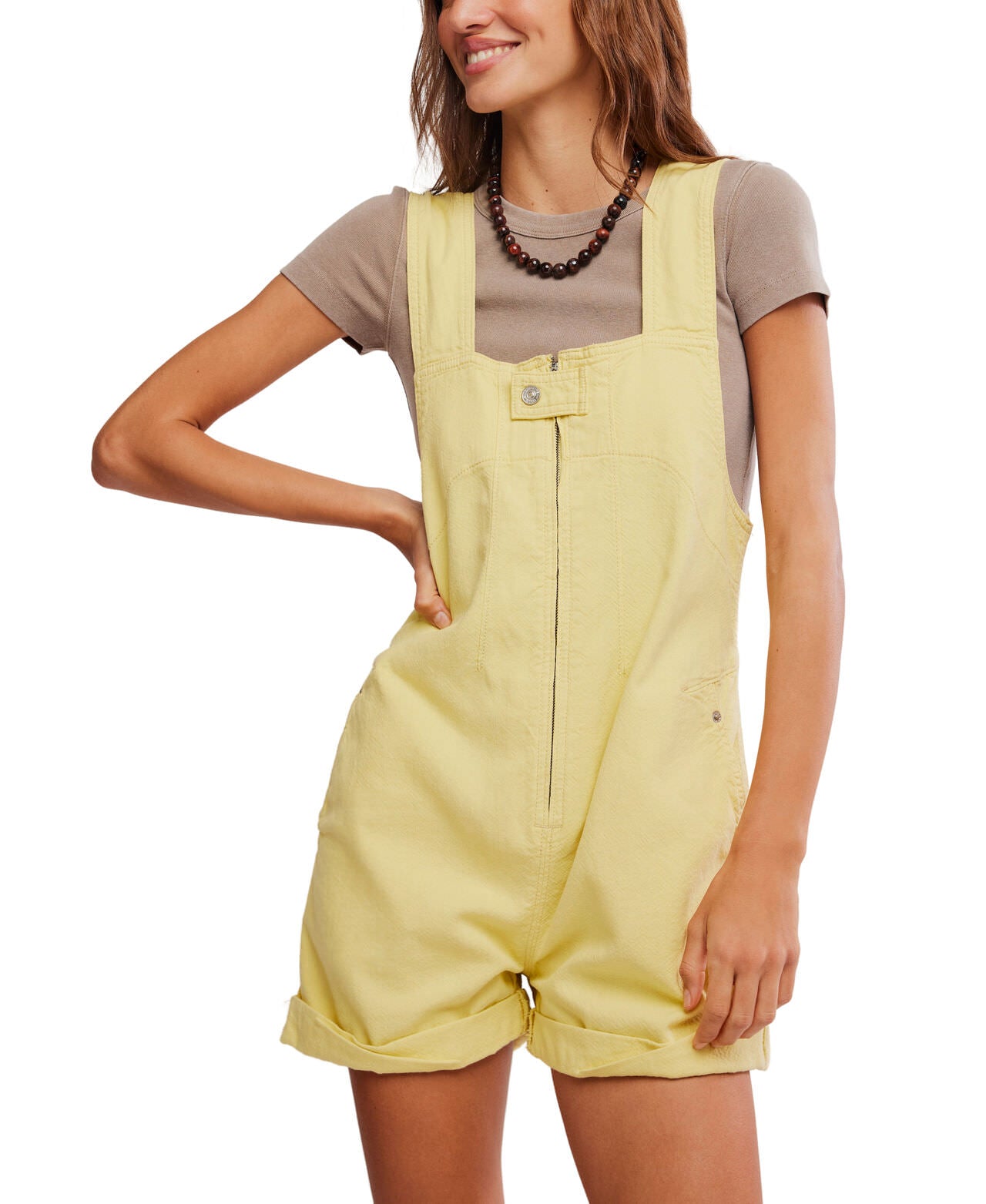 A woman smiles slightly, posing with one hand on her hip and wearing the Free People Jude Shortall in yellow over a beige short-sleeve t-shirt, accessorized with a beaded necklace.