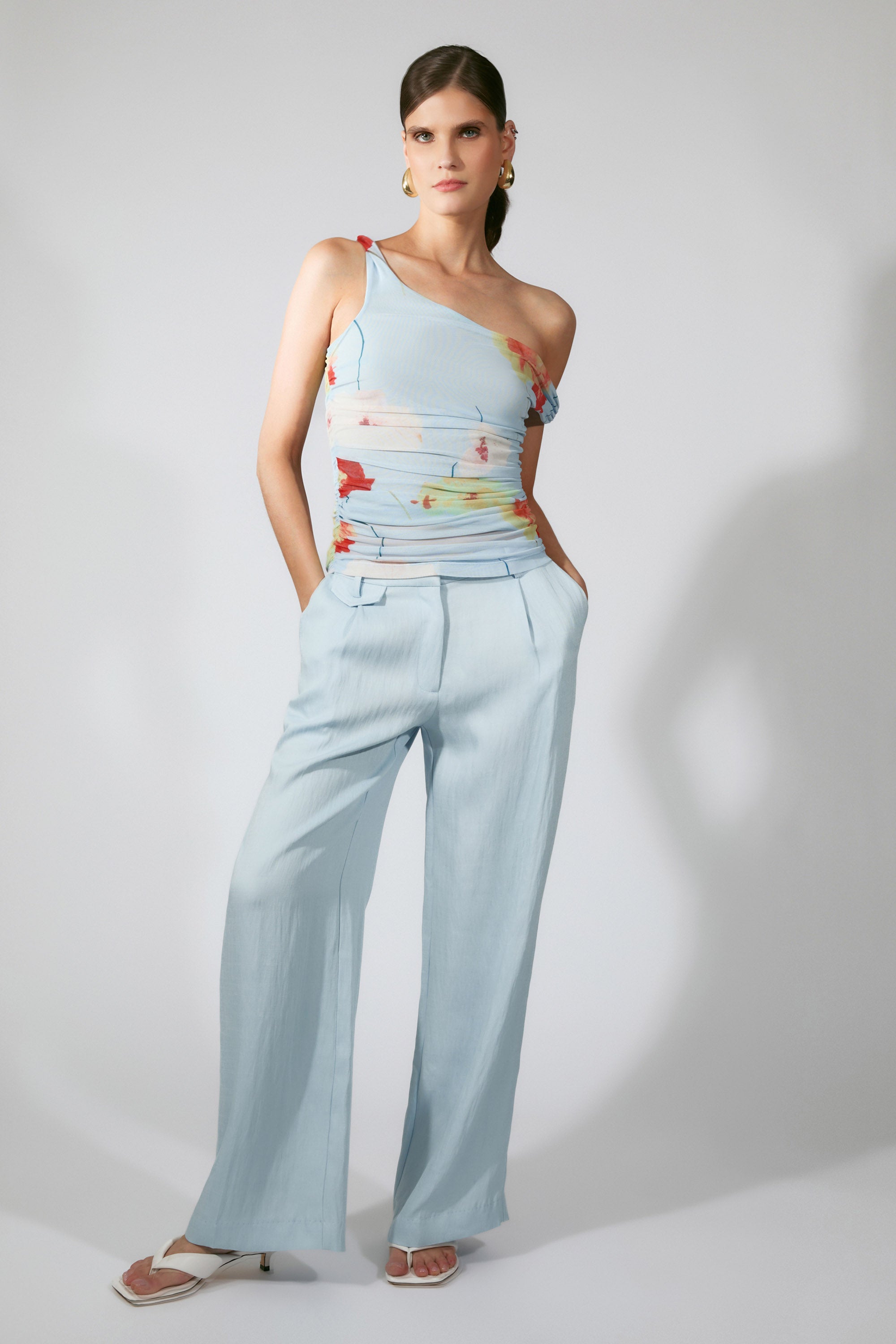 A woman stands against a plain background, wearing light blue wide-leg trousers and the Bardot Layana Printed Mesh Asym Top with a pastel floral pattern. She pairs it with white heeled sandals, gold earrings, and has her hands in her pockets.