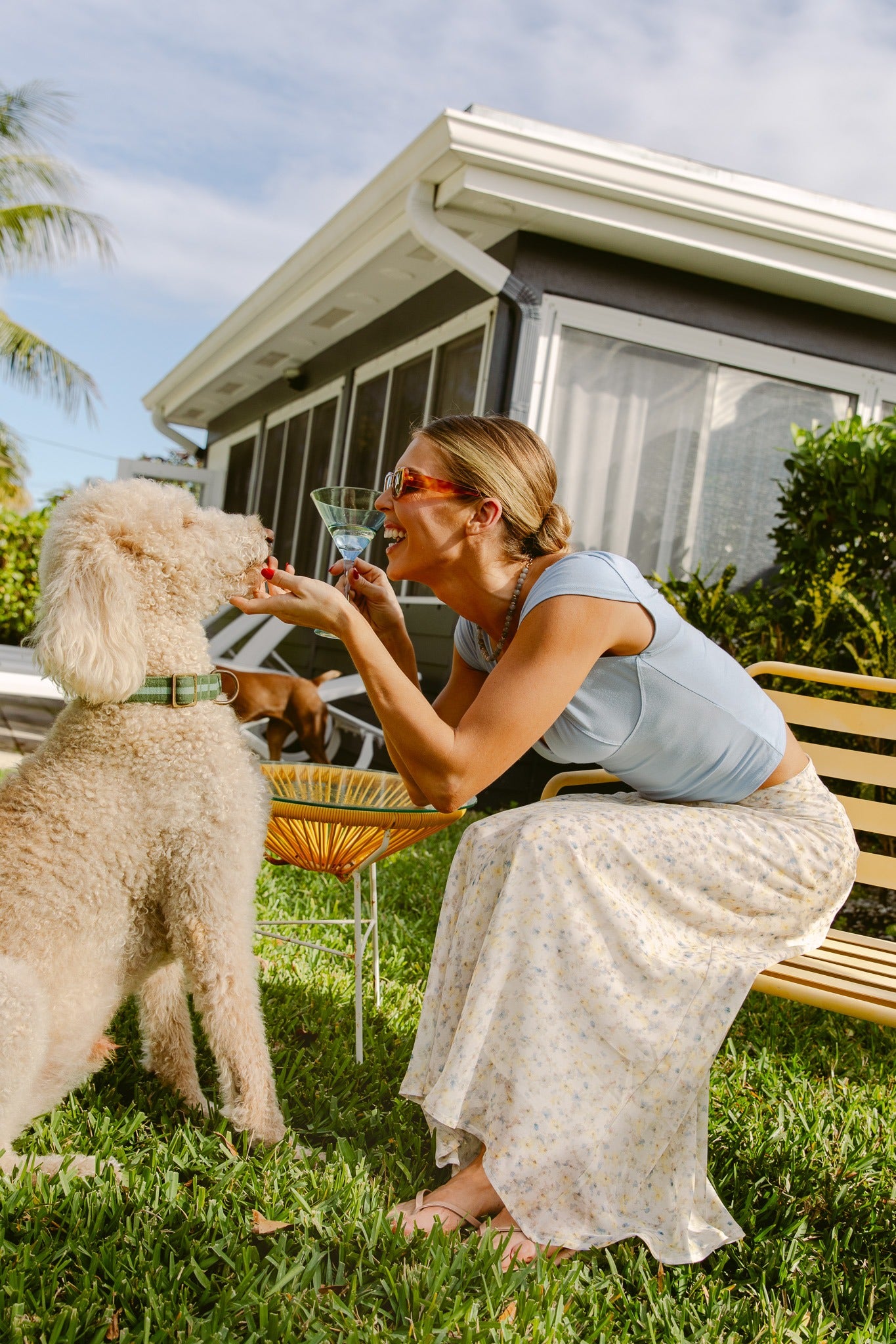 A woman in a blue top and the South Moon Under Mollie Floral Midi Skirt sits on a bench outside, smiling as she holds up a treat for a large, curly-haired dog. A house and greenery are visible in the background.