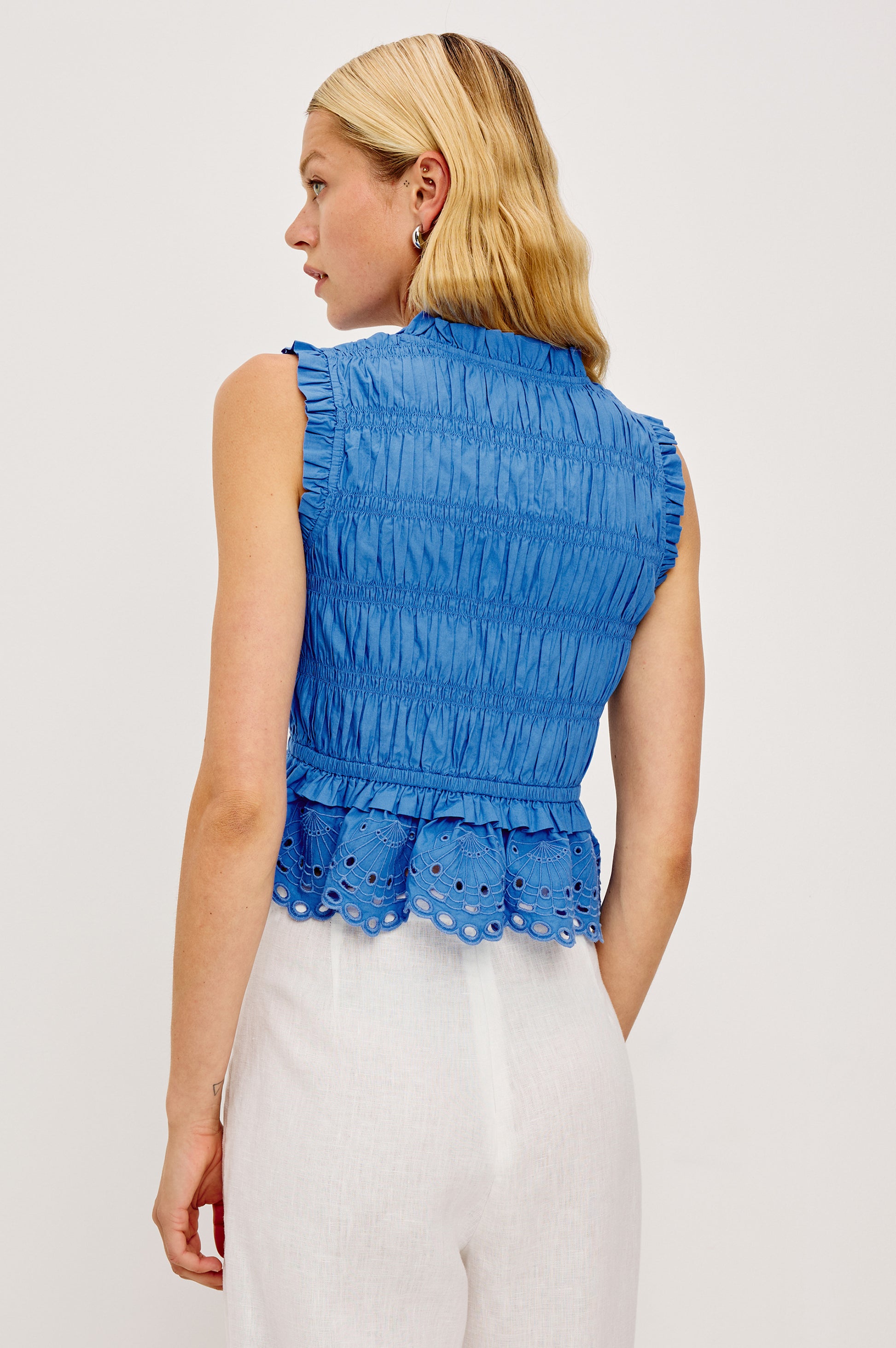 A woman with blonde hair wears the Rails Martine Sleeveless Top in blue cotton poplin, featuring ruffle details, paired with white high-waisted pants. She faces away and looks to the side against a plain background.