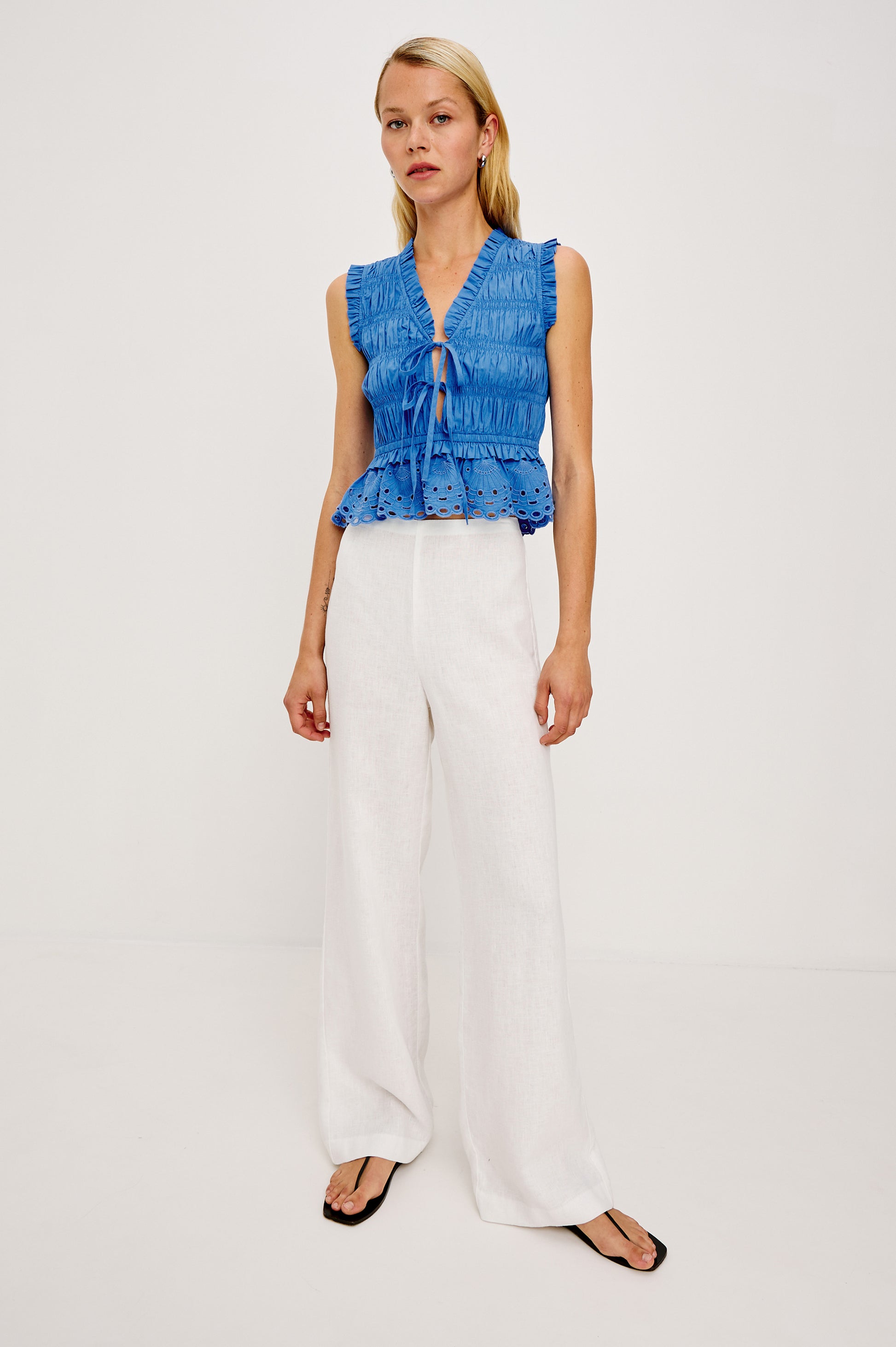 A woman with straight blonde hair and a neutral expression wears the Rails Martine Sleeveless Top, featuring blue cotton poplin with ruffles and a tie front, paired with white wide-leg pants and black sandals against a plain background.