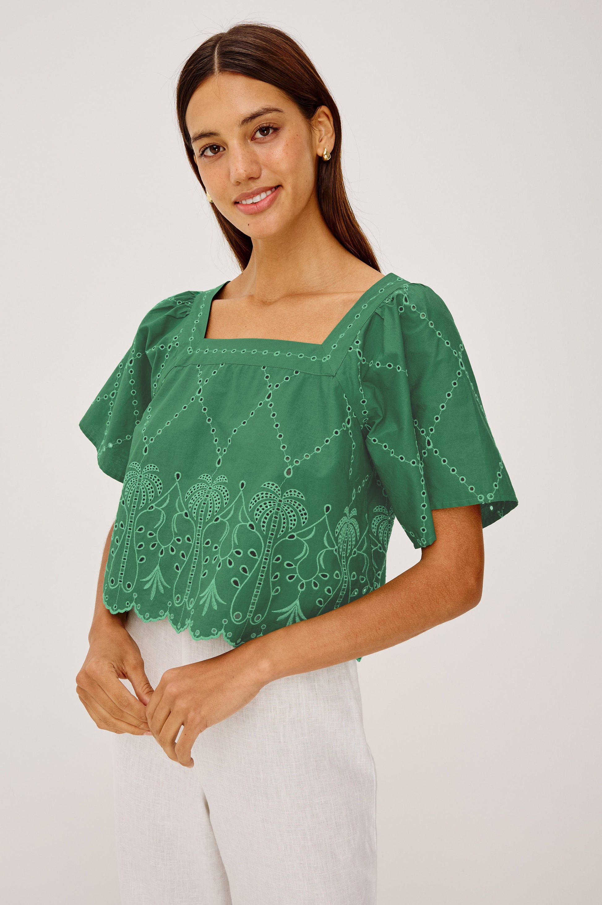 A woman smiles in the Rails Laine top, a green short-sleeved cotton poplin piece with embroidered patterns, paired with white pants against a plain light background.