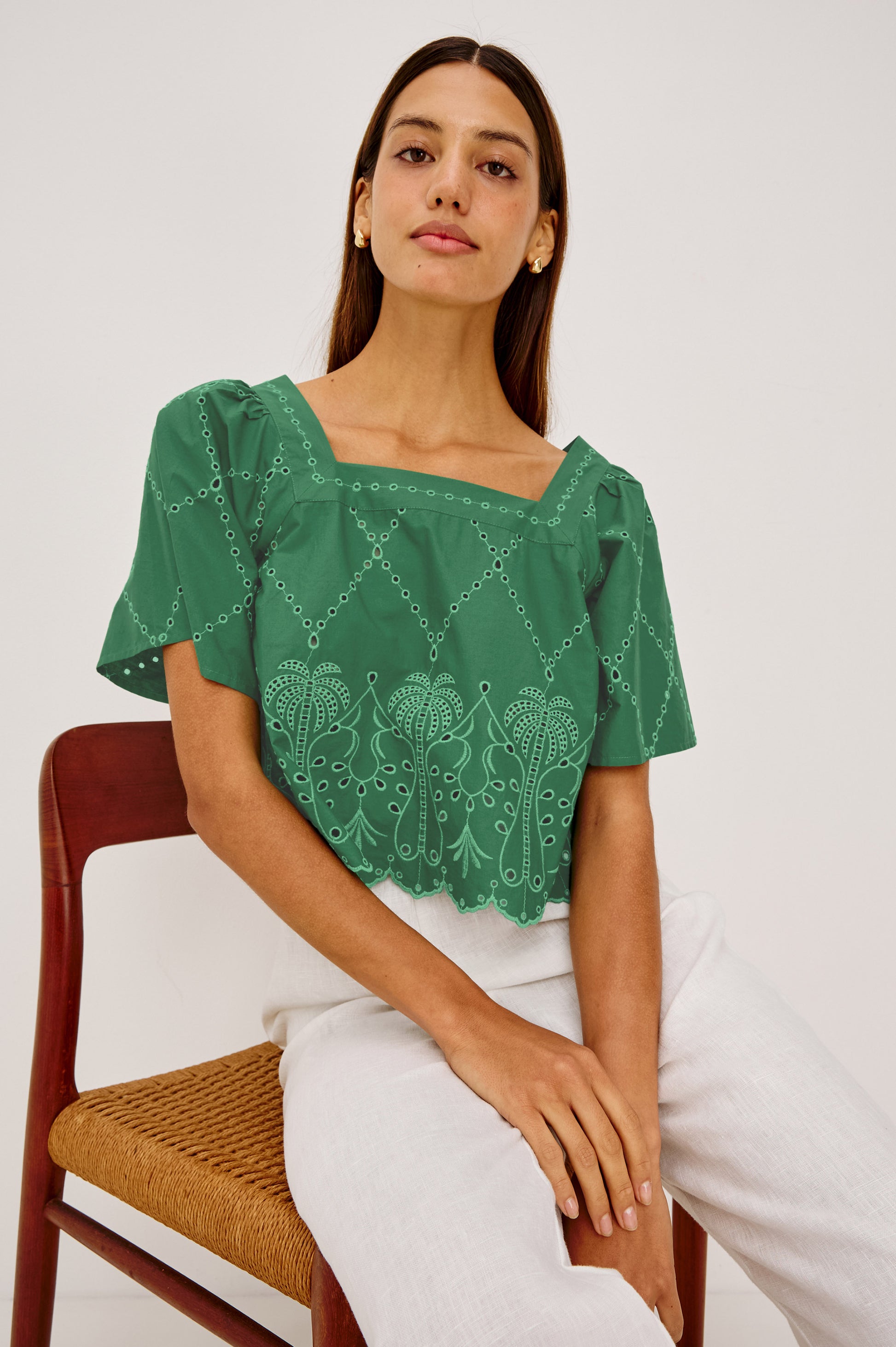 A woman with straight brown hair sits on a wooden chair, confidently wearing the Rails Laine green cotton poplin top with embroidered details and white pants, against a plain background.