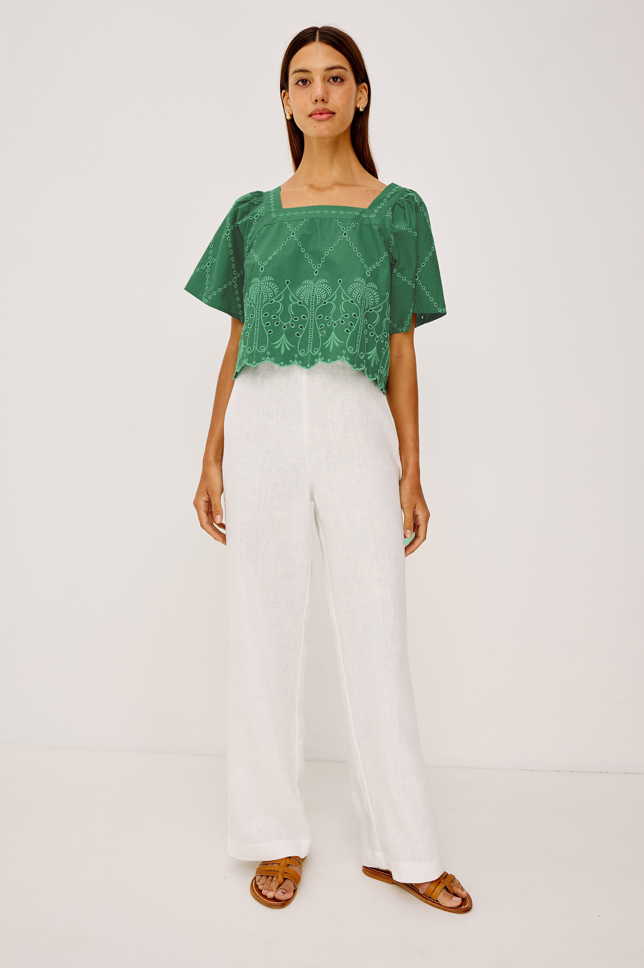 A woman stands against a plain background wearing the Rails Laine green short-sleeve square-neck blouse with embroidered patterns and a scalloped hem, white wide-leg pants, and brown sandals.