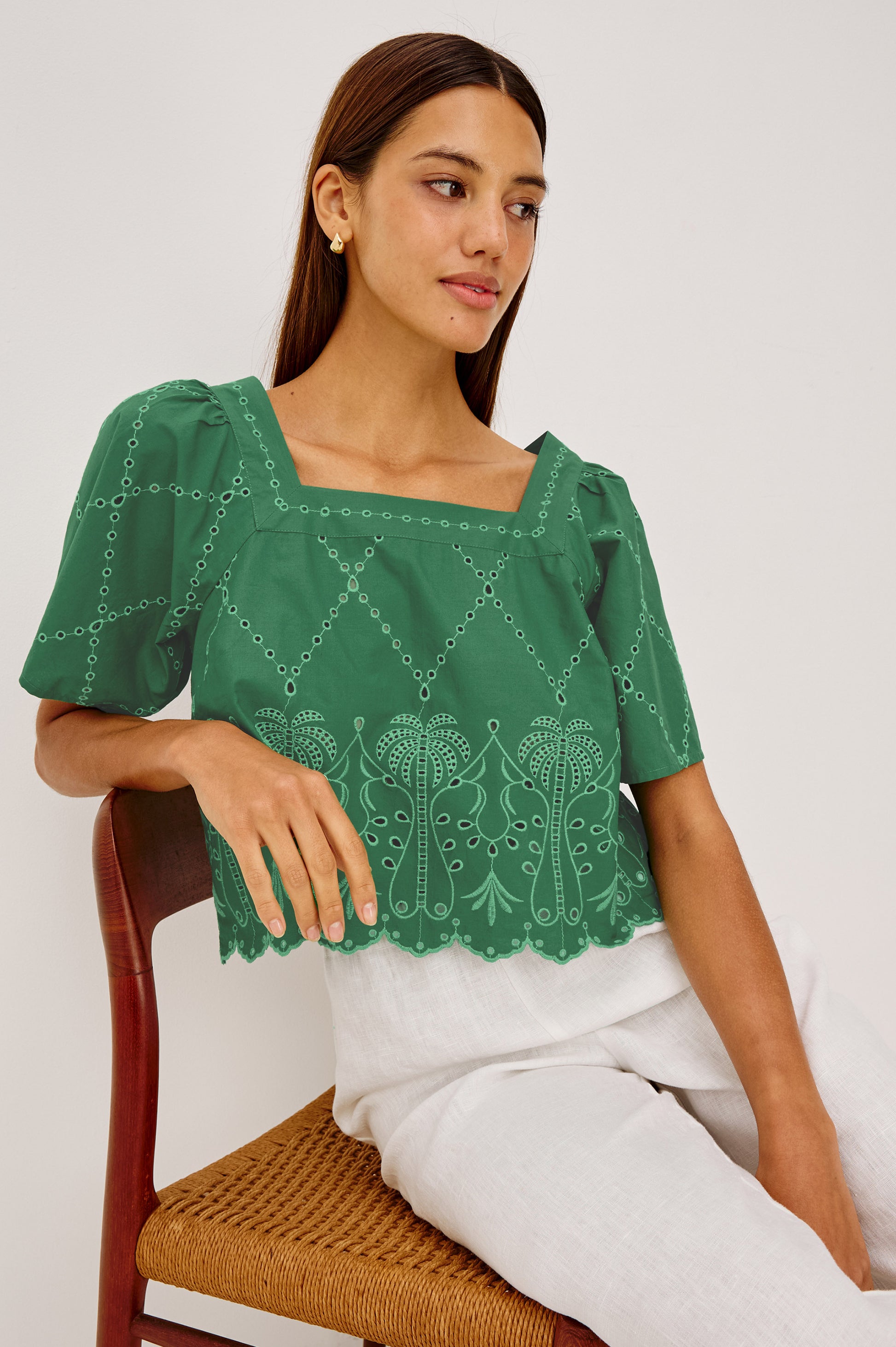 A woman with straight brown hair sits on a chair, wearing the Rails "Laine" top in Green Multi, featuring embroidered patterns and a scalloped hem, paired with white pants. She gazes to the side with a neutral expression against a plain background.
