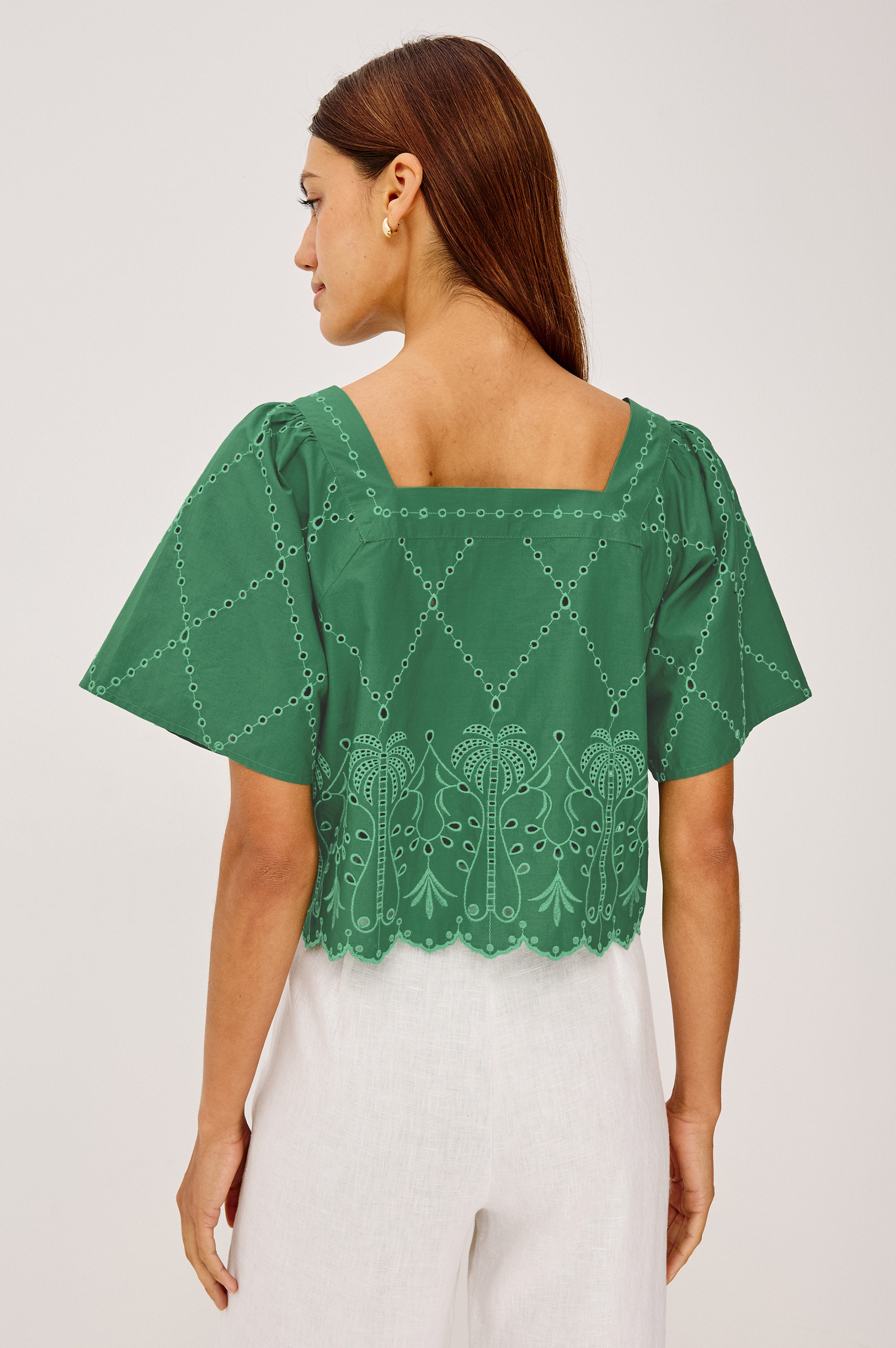 A woman with straight brown hair wears the Rails Laine green embroidered cropped cotton top with a square neckline and scalloped hem, paired with high-waisted white pants, standing with her back to the camera.