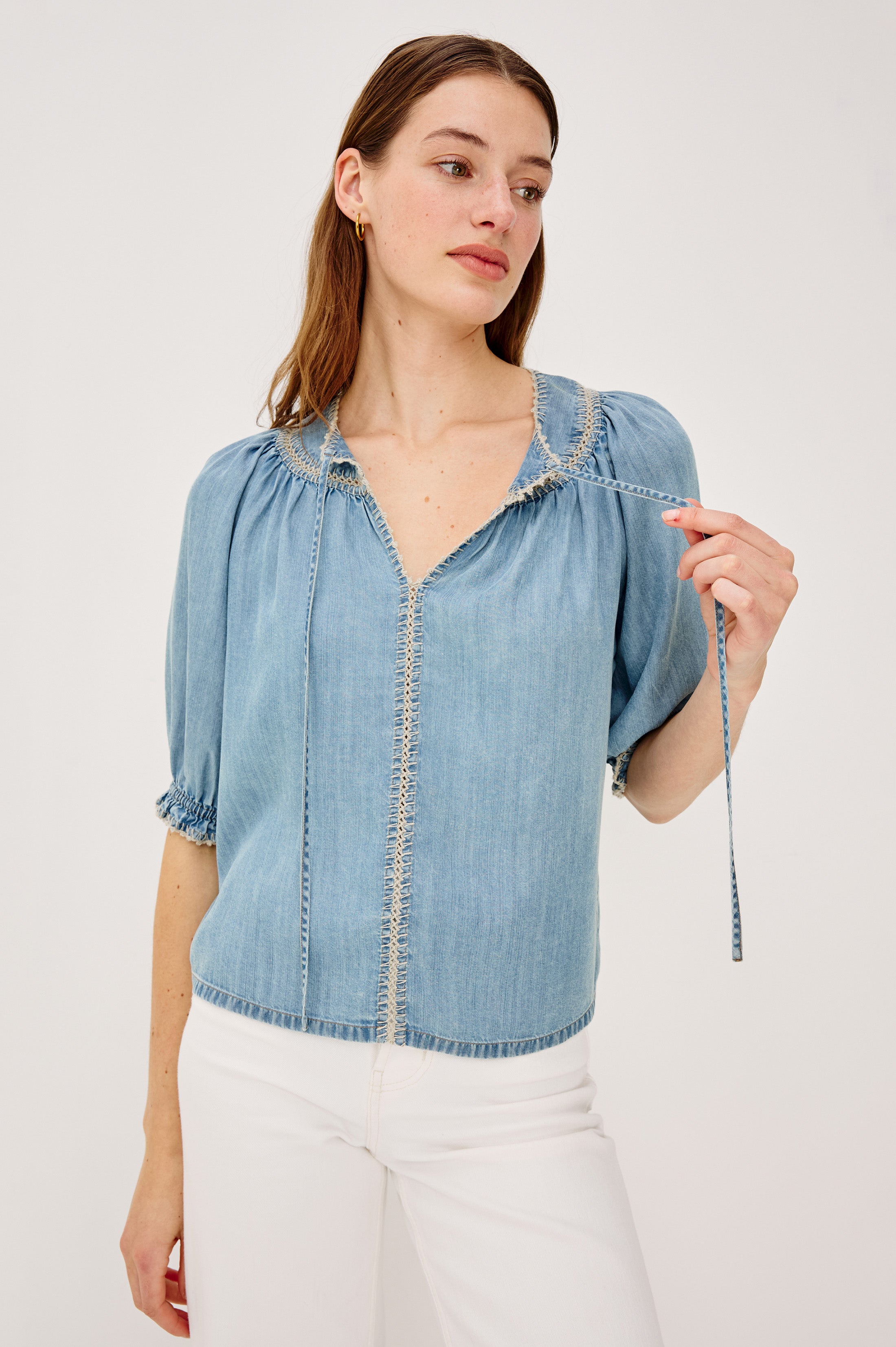 A woman models the Rails Belina Short Sleeve Denim Top in light blue TENCEL with decorative stitching and a peasant-style V-neckline, paired with white pants, against a plain white background while holding one of the blouse’s ties.
