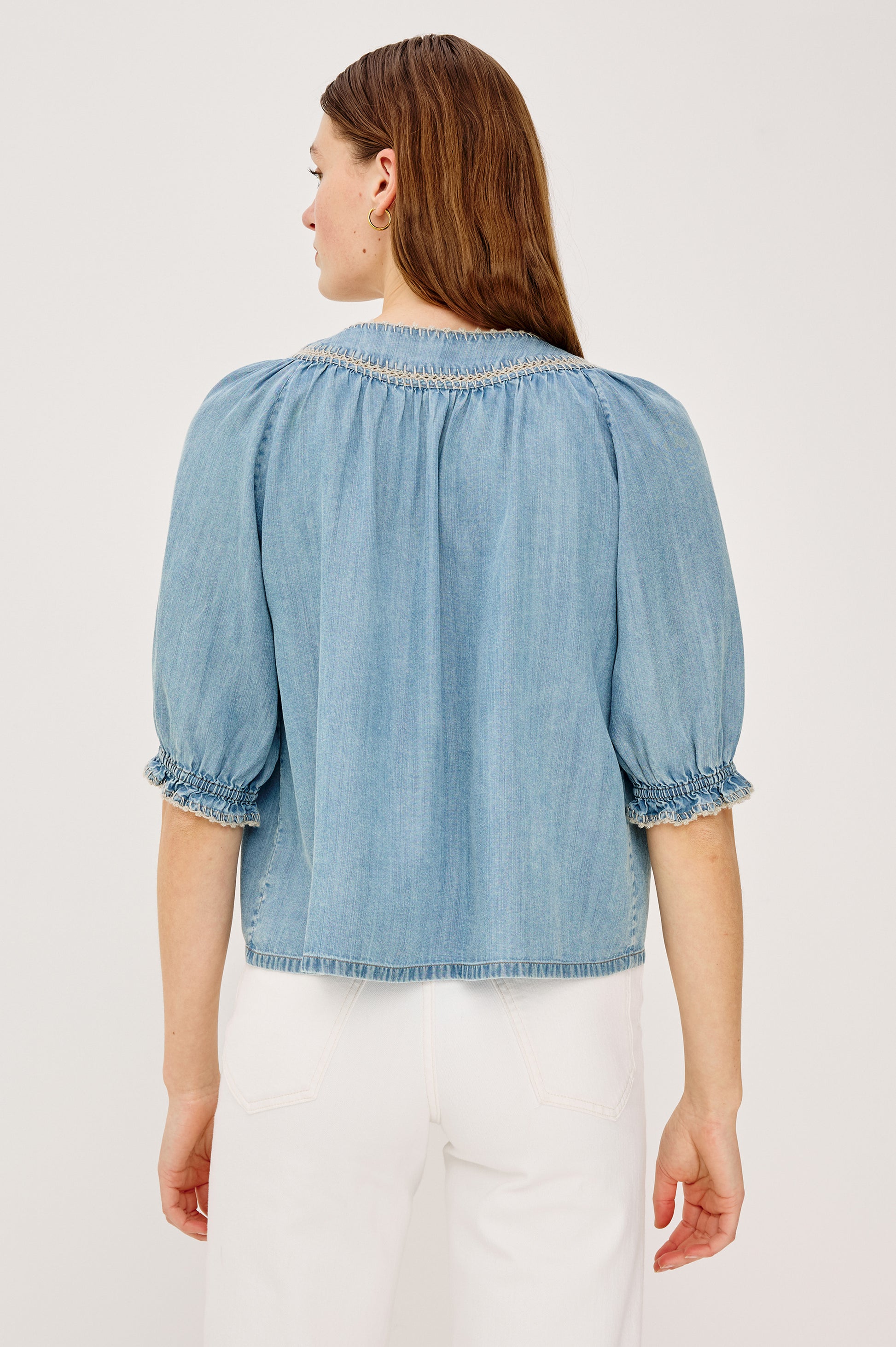 A woman faces away, wearing the Rails Belina Short Sleeve Denim Top with shirred cuffs and neckline, paired with white pants. Her straight brown hair falls loosely down her back.