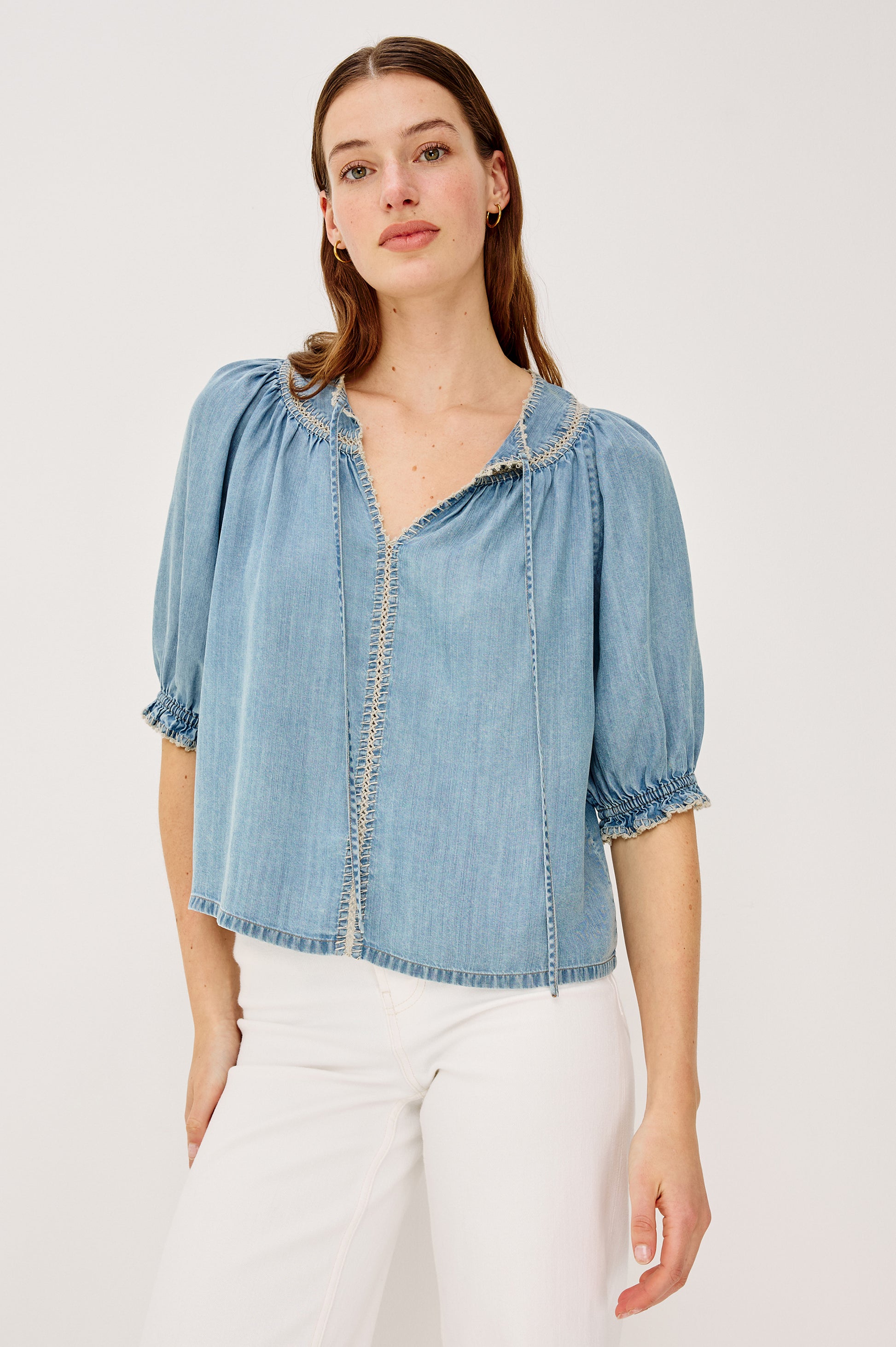 A woman wears the Rails Belina Short Sleeve Denim Top, featuring ultra-soft TENCEL fabric, puffed half sleeves, a peasant-style V-neckline, and decorative trim, paired with high-waisted white pants against a plain light background.