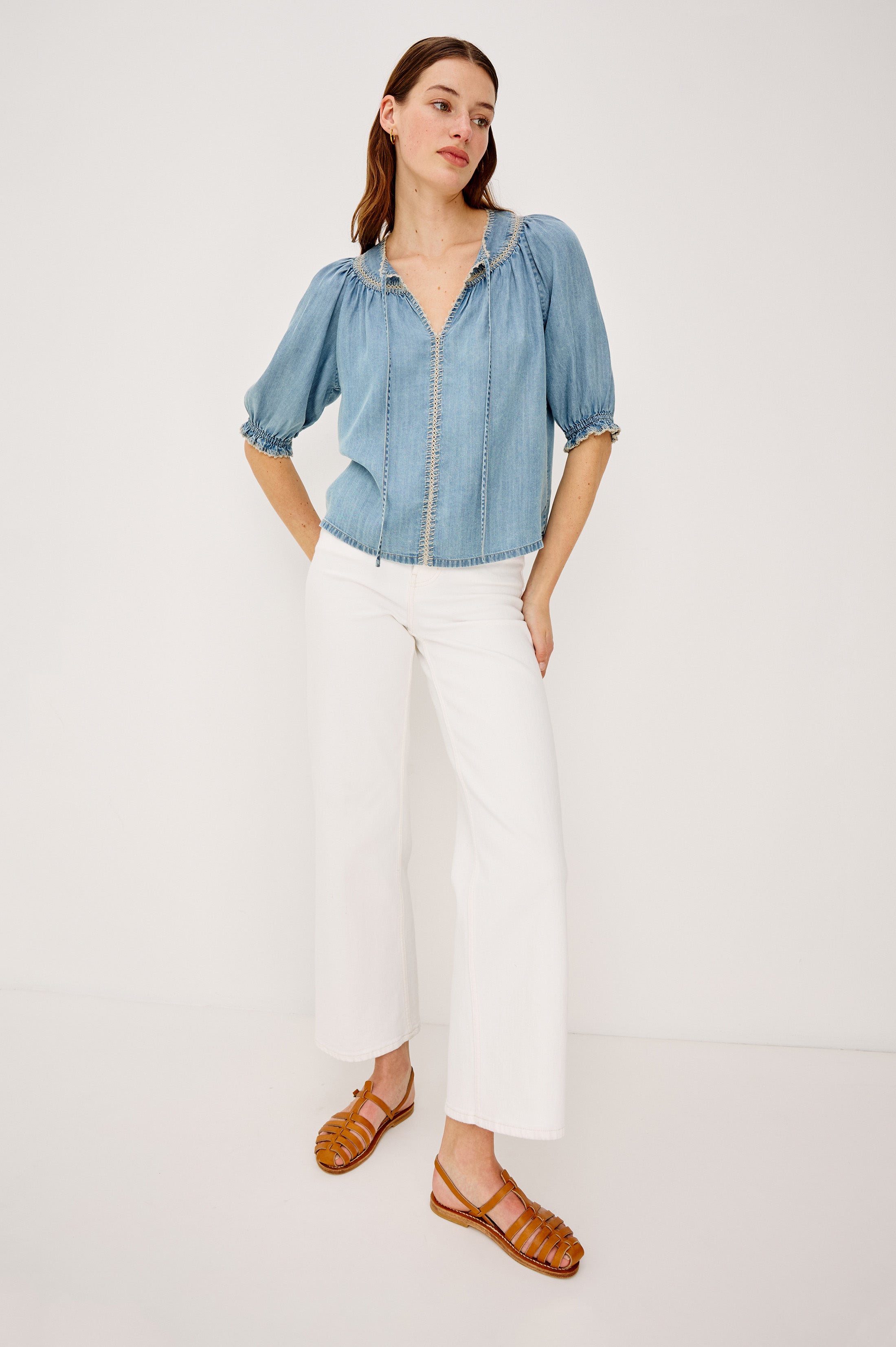 A woman with straight brown hair stands against a white background wearing the Rails Belina Short Sleeve Denim Top, featuring puff sleeves and a peasant-style V-neckline. She pairs it with white wide-leg pants and brown strappy sandals, hands in her pockets.