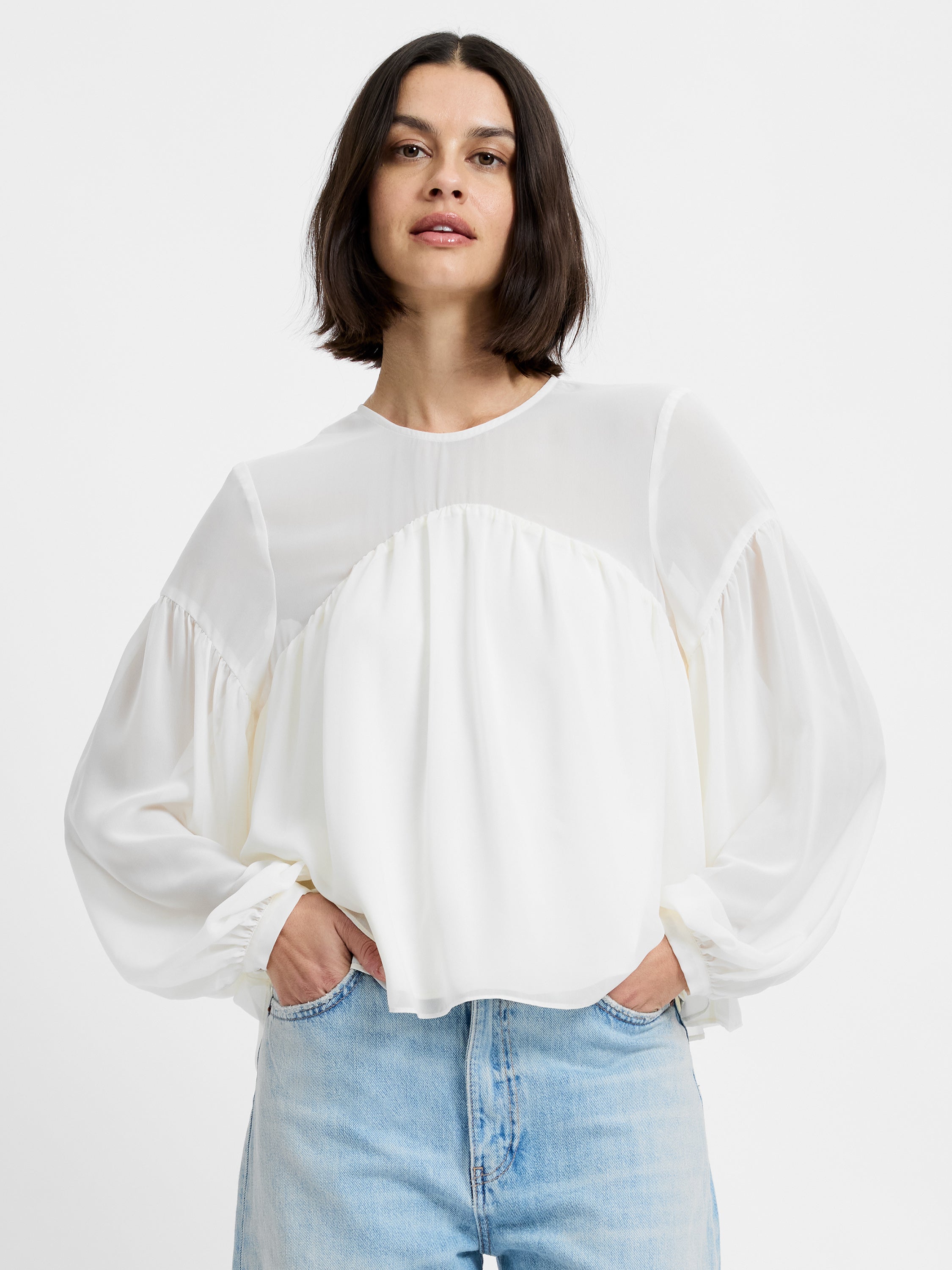 A woman with a bob haircut wears the French Connection Marina Silky Curved Yoke blouse in cream paired with light blue jeans, standing with her hands in her pockets against a plain white background.
