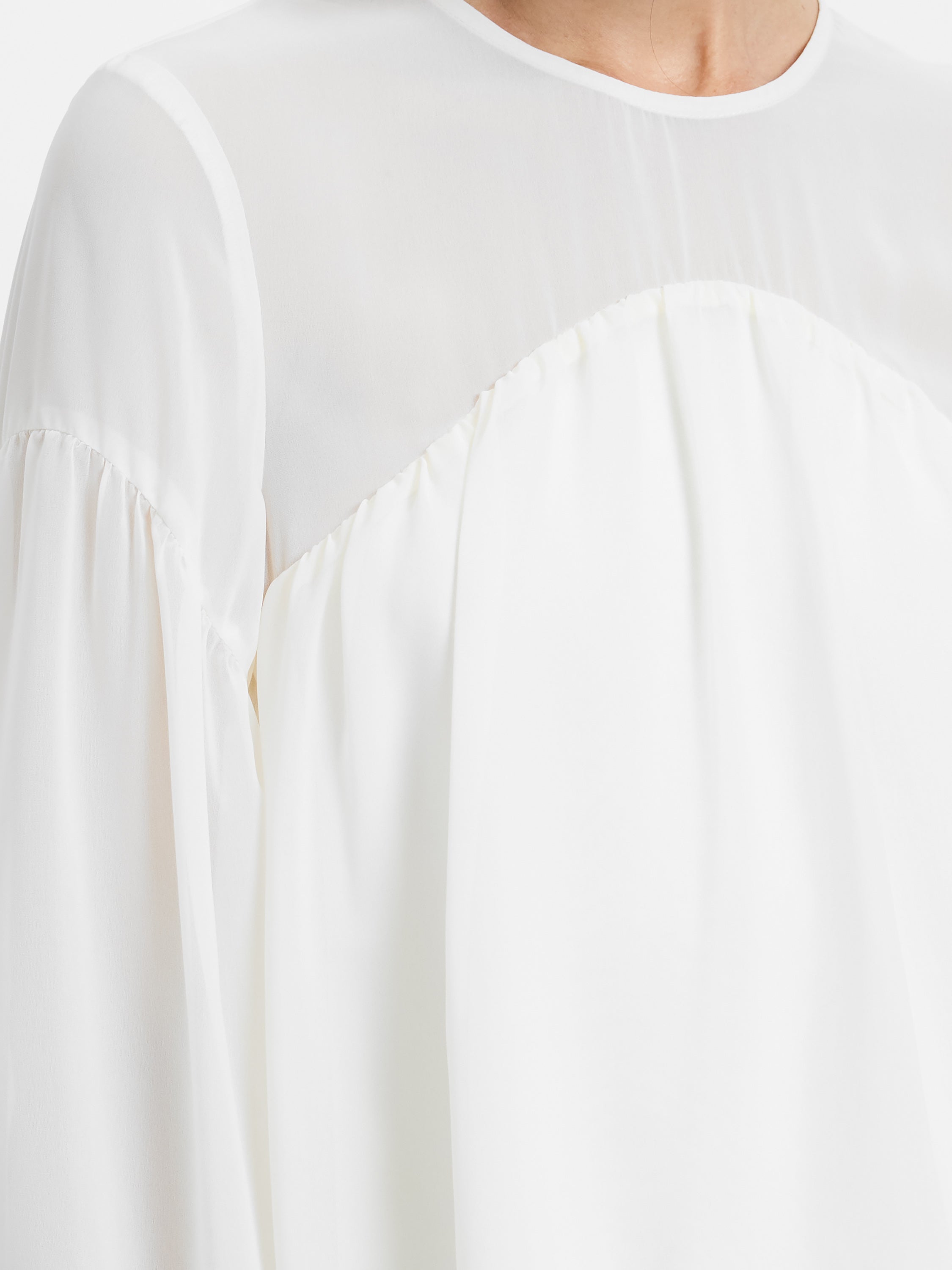 A person wears the French Connection Marina Silky Curved Yoke—a white, long-sleeved lightweight blouse with sheer sleeves and upper chest, plus gathered detailing below the bust.