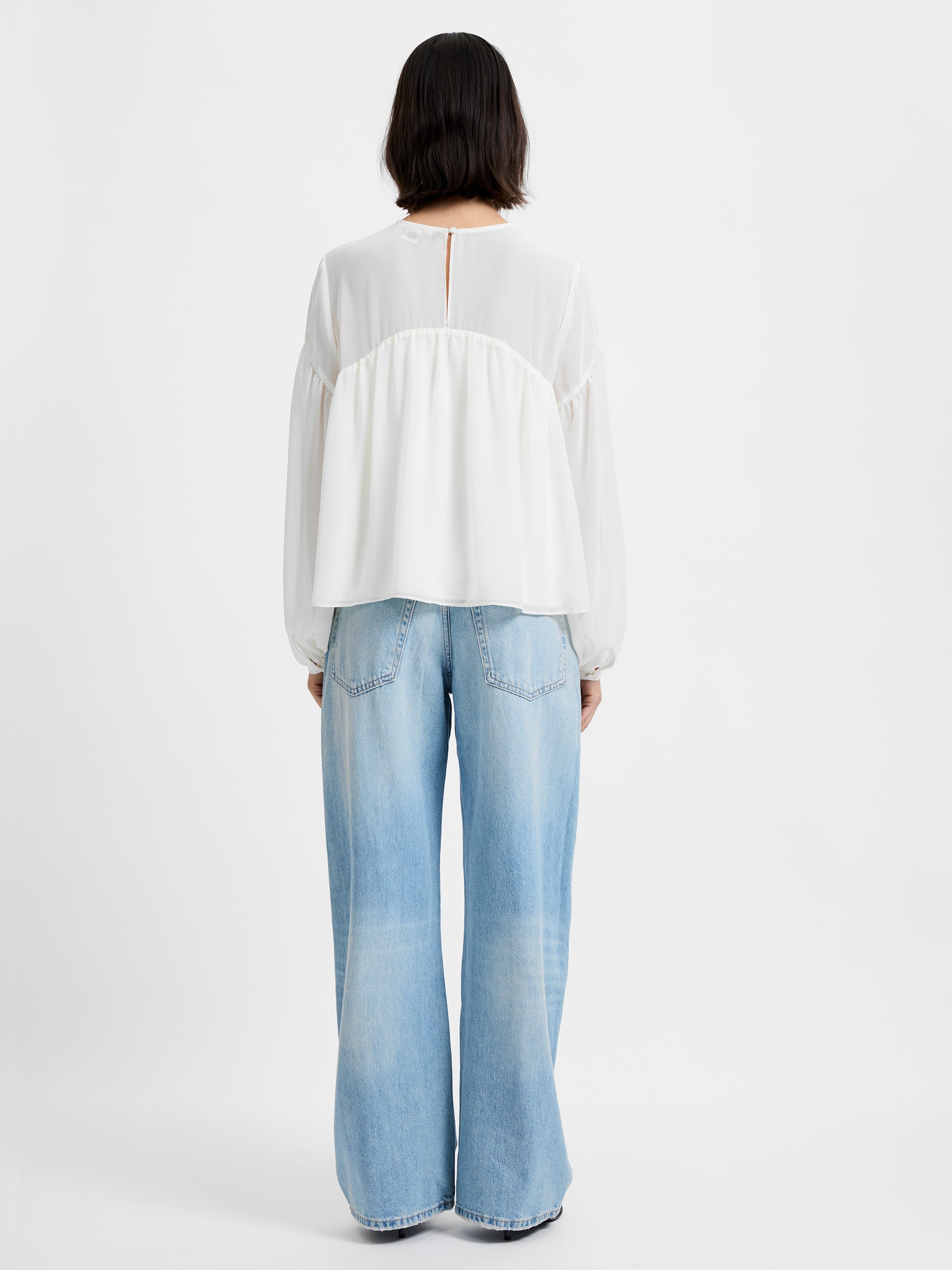 A person faces away from the camera, wearing the French Connection Marina Silky Curved Yoke blouse and light blue wide-leg jeans, posed in front of a plain white background.