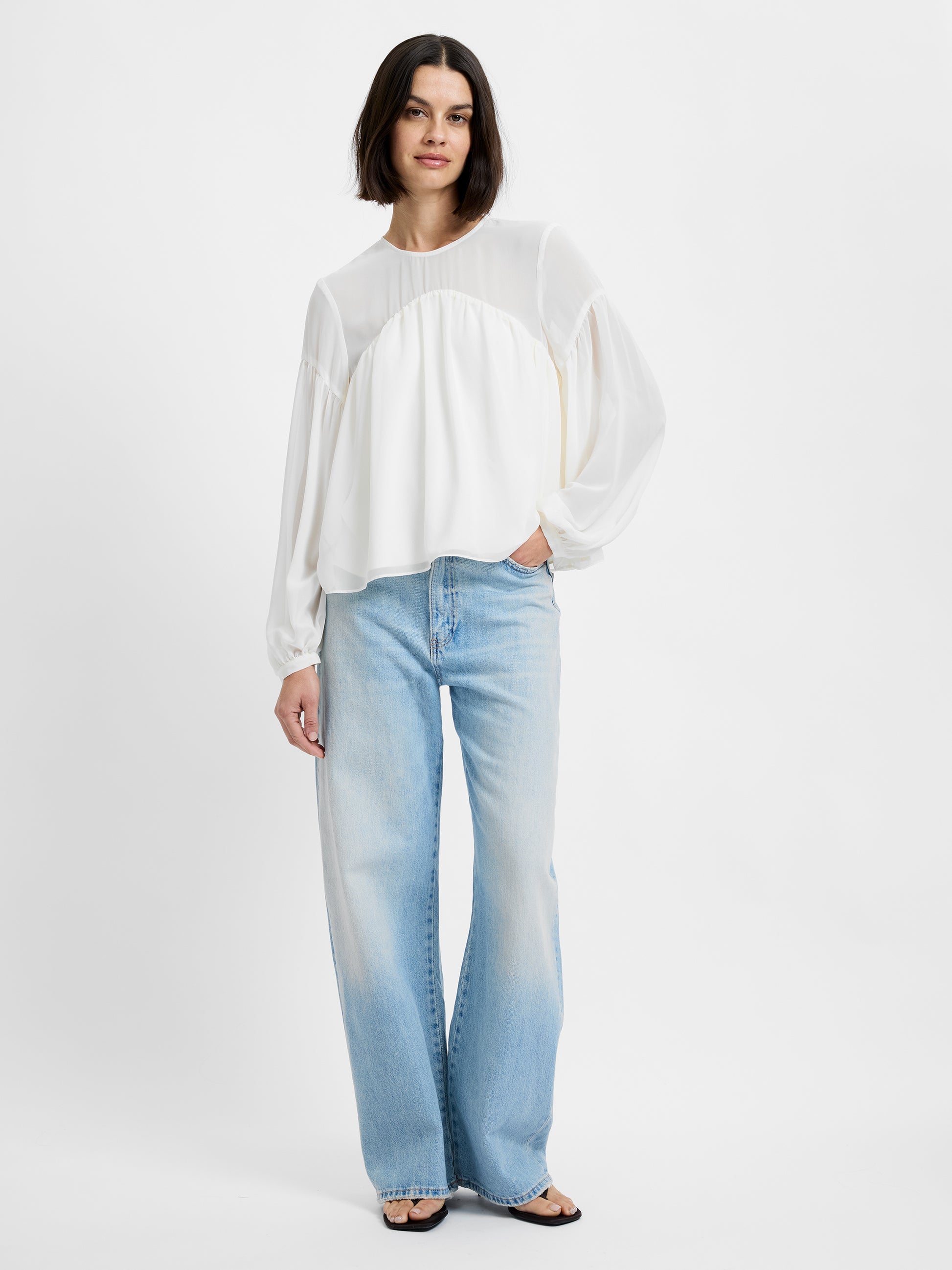 A woman wears the French Connection Marina Silky Curved Yoke blouse, styled with light-wash wide-leg jeans and black sandals. She stands against a plain white background with one hand in her pocket and a neutral expression.