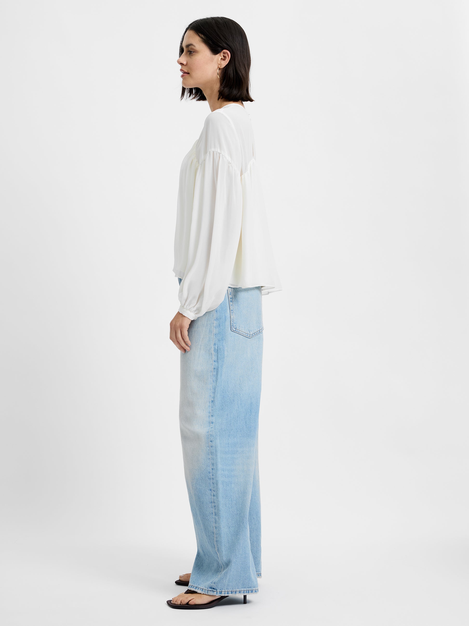 A woman stands in profile against a white background, wearing the French Connection Marina Silky Curved Yoke blouse, light blue wide-leg jeans, and black open-toe sandals.