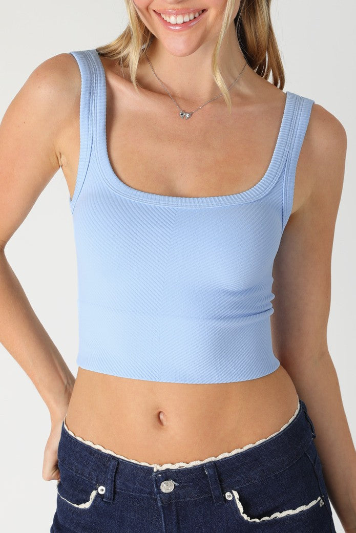 A woman with blonde hair down, smiling, wears the Niki Biki Chevron Rib Crop Top in SERENE BLUE and dark denim jeans with white scalloped trim. Only her upper body is visible.