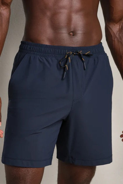 A man wears RHONE Pursuit Shorts 7 In Unlined in navy, featuring an elastic waistband, drawstring, side pockets, and a relaxed standard fit. The shorts are shown from torso to upper legs, with his hands resting at his sides.