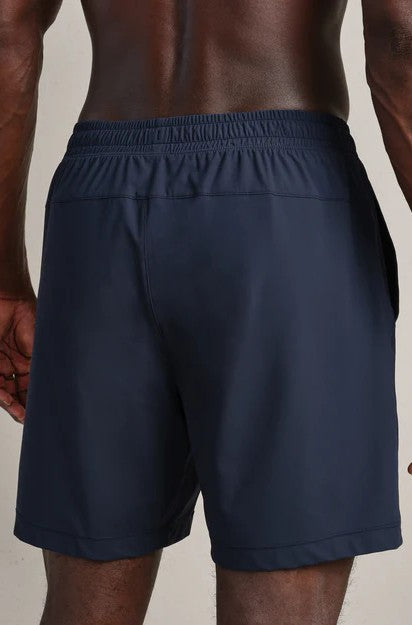 A person is seen from behind wearing RHONE Pursuit Shorts 7 In Unlined in navy blue, featuring a 7" inseam and elastic waistband. The neutral background highlights the back and upper legs of the wearer.