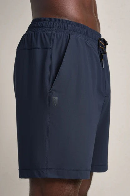 A person wears RHONE Pursuit Shorts 7 In Unlined, featuring an elastic waistband, side pockets, and a dark blue color, shown from the side against a plain light background.