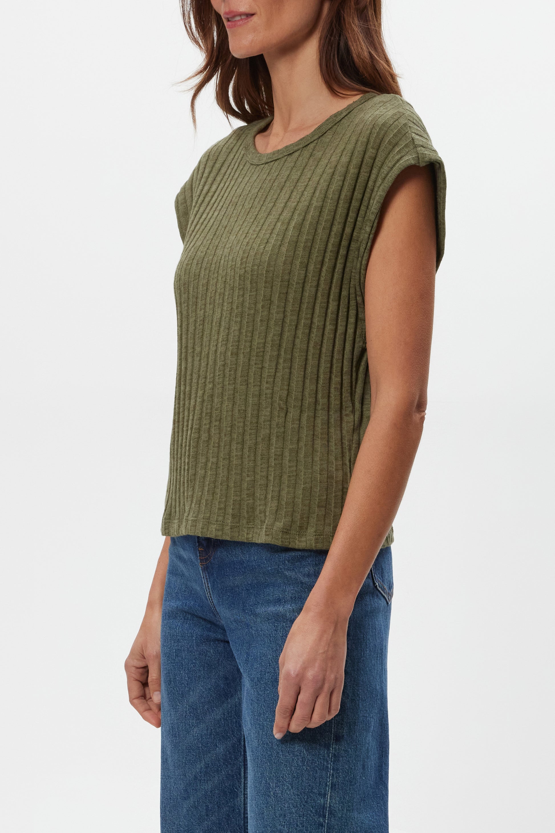 A woman models the Michael Stars Jinny Power Shoulder Top in olive green, a ribbed, fitted cotton-spandex style with short sleeves. She pairs it with blue jeans and stands against a white background, her head partially out of frame.