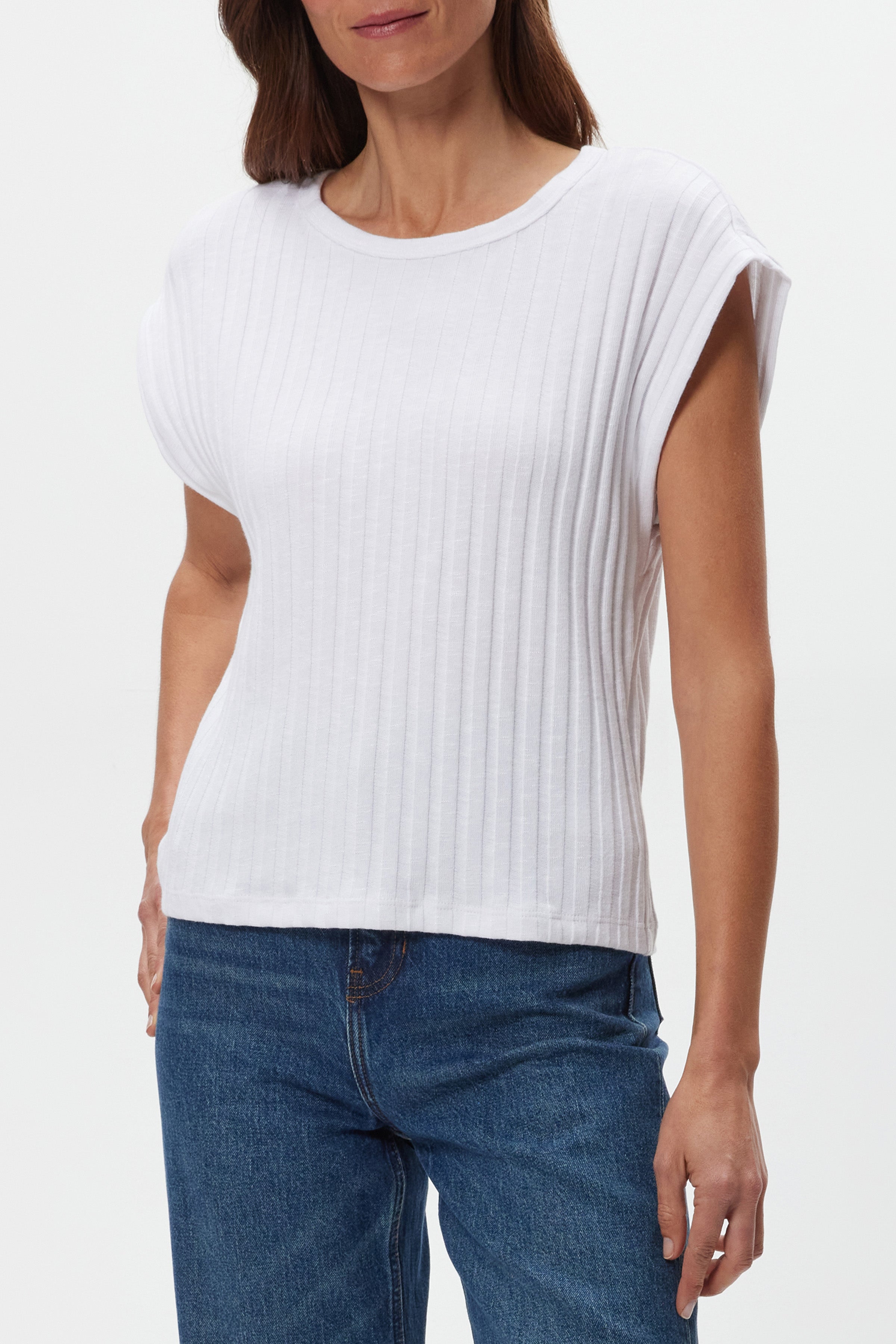 A woman models the Michael Stars Jinny Power Shoulder Top in white, featuring short sleeves and a fitted cotton-spandex blend. She pairs it with blue jeans, standing against a plain white background with her head partially cropped.