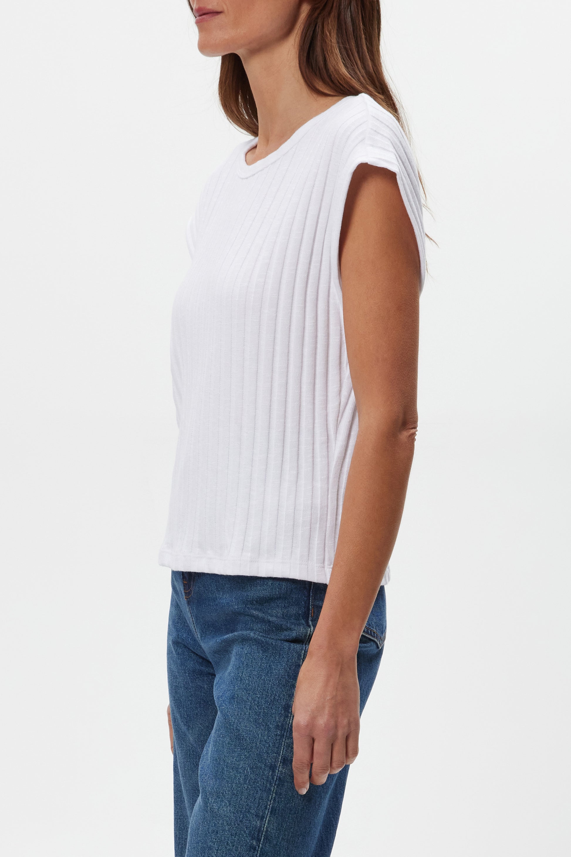 A woman is shown in profile wearing the white, sleeveless Jinny Power Shoulder Top by Michael Stars with blue jeans against a plain white background.