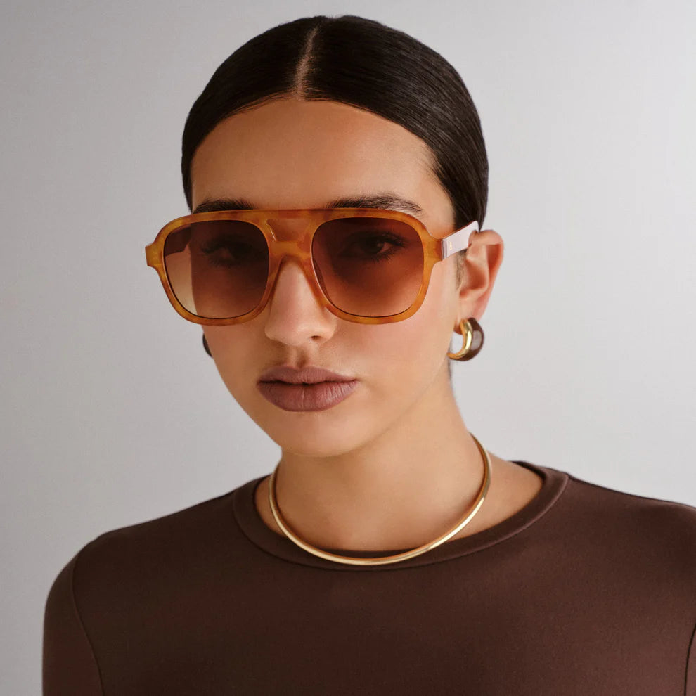 A person with slicked-back dark hair wears Aire’s Whirlpool Sunnies—large sunglasses with brown gradient lenses and a vintage tort frame—plus gold hoop earrings, a gold necklace, and a brown top against a plain light background.