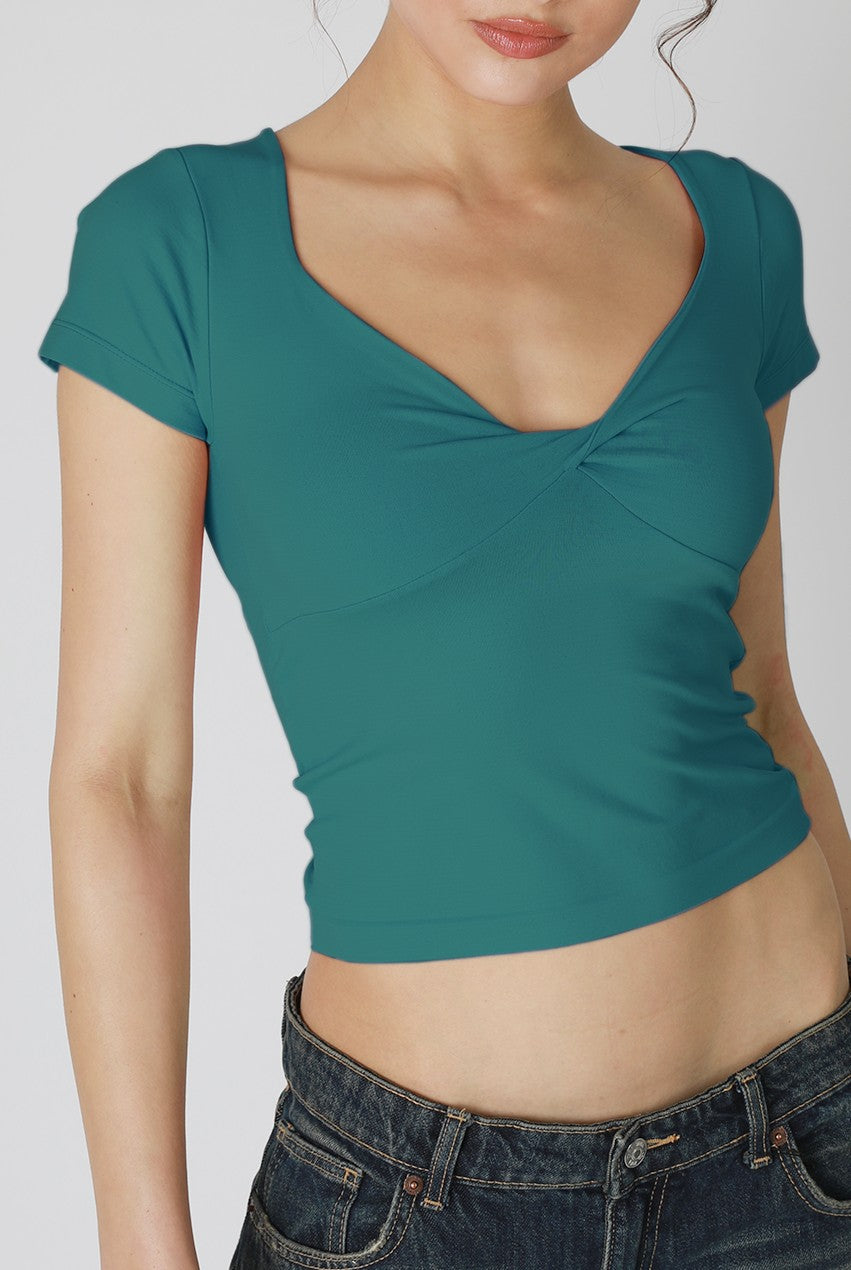 A woman wears a dark green Niki Biki Double Twist Short Sleeve tee with blue jeans, standing against a plain light background. Her head is slightly turned and partially out of frame—a versatile staple for everyday wear.