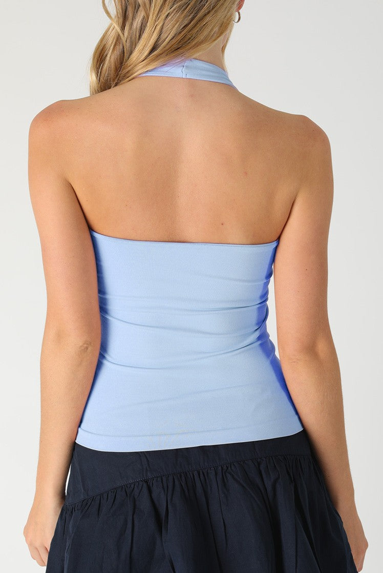 A woman is seen from behind in a Niki Biki Smooth Halter, light blue, and a dark blue skirt. Her blonde hair rests over one shoulder, set against a plain background, capturing effortless style and everyday comfort.
