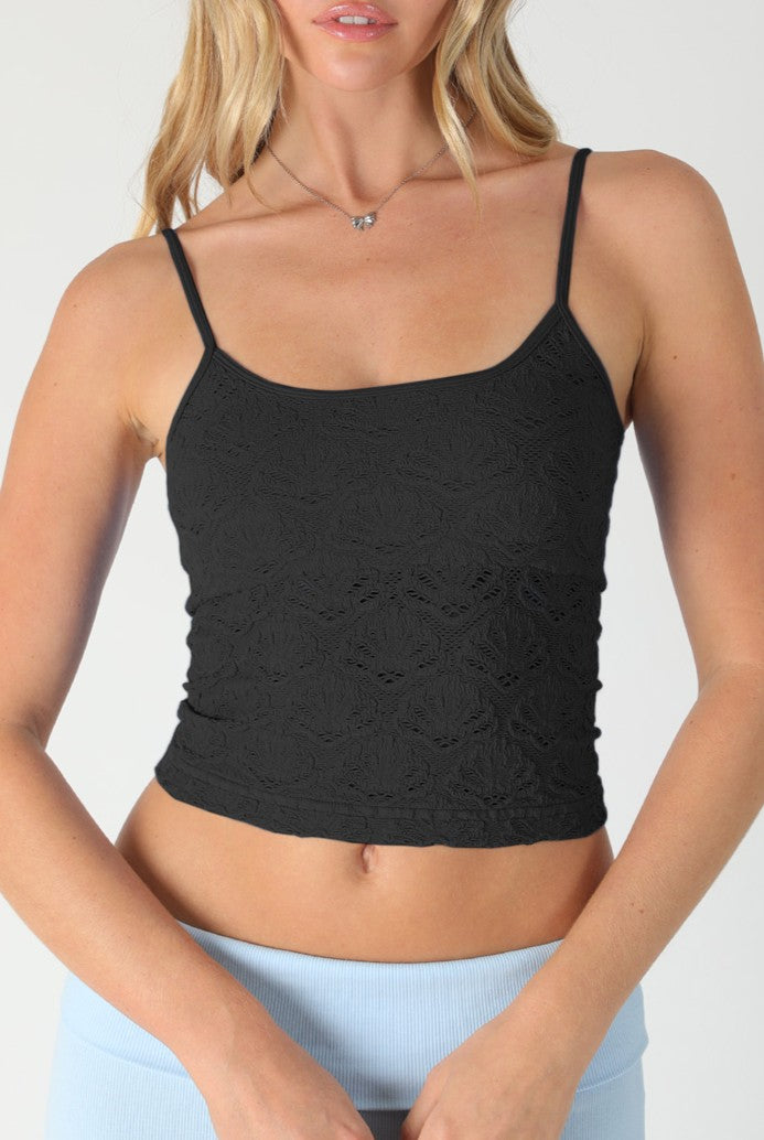 A woman wears the Niki Biki Floral Lace Textured Cami, a sleeveless black crop top with thin straps and seamless fit, paired with light blue pants. She stands against a plain light background, her face partially out of frame.