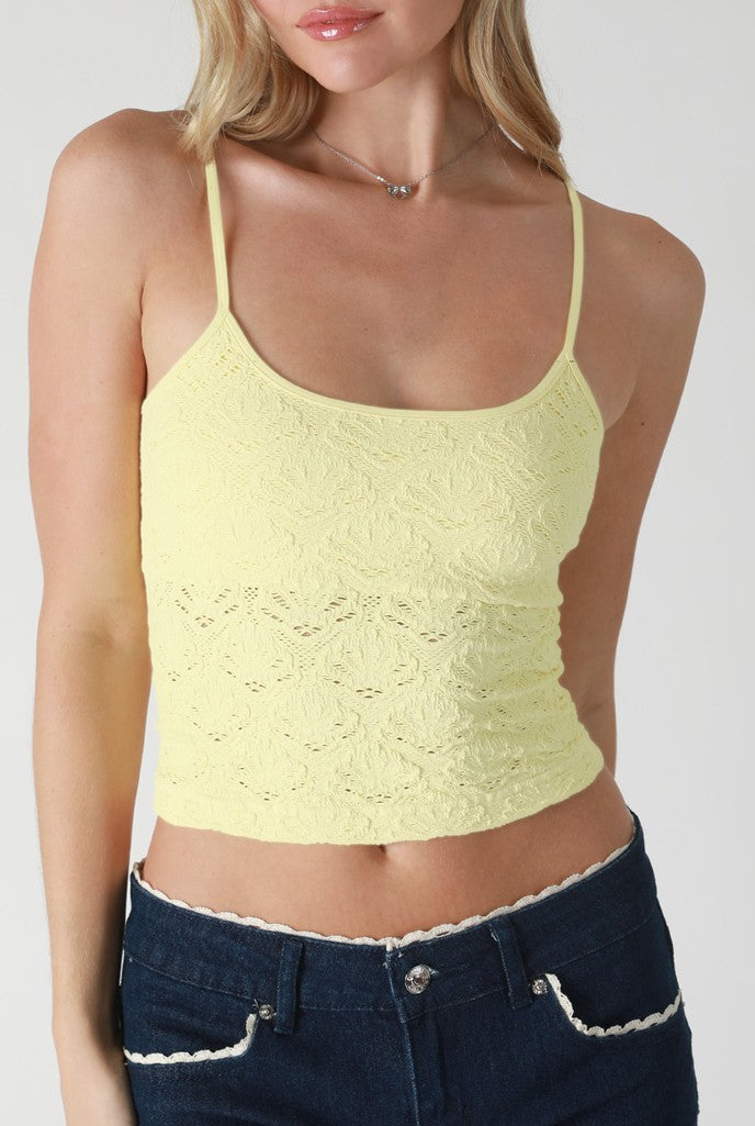 A woman wears the Niki Biki Floral Lace Textured Cami in light yellow with thin straps and dark blue high-waisted jeans. The image, cropped shoulder to hip, highlights the cami's seamless fit ideal for comfortable layering.