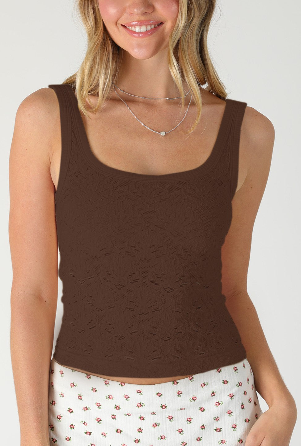 Smiling woman wearing the Niki Biki Floral Textured Lace Tank in COFFEE BEAN and a white skirt with a red and green floral pattern, standing against a plain light background.