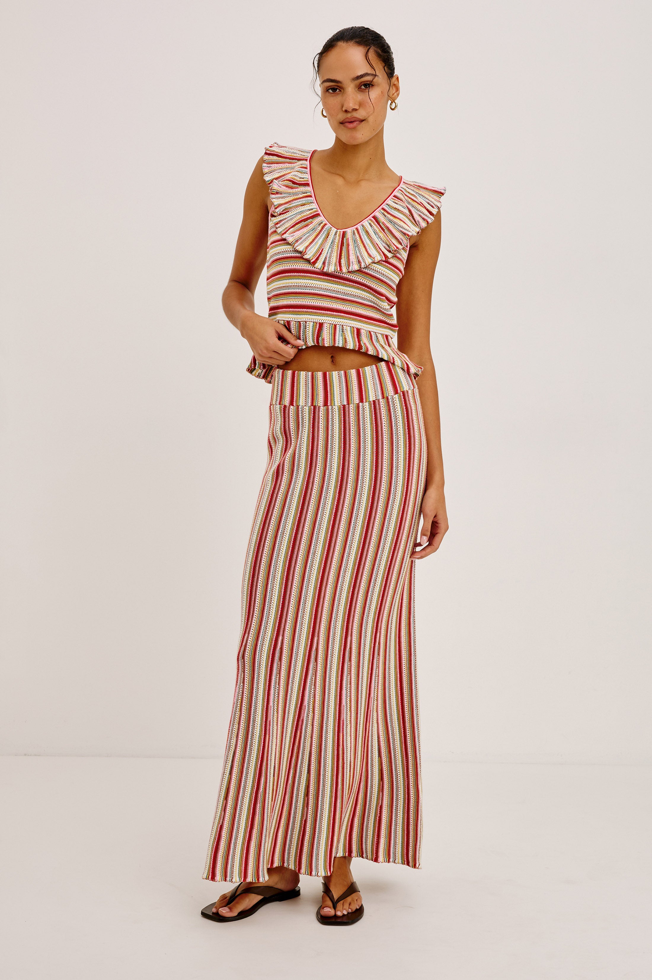 A woman stands barefoot on a white background wearing the Rails Nari Skirt, a high-waisted maxi skirt with vertical red, white, and beige stripes, paired with a sleeveless ruffled crop top.