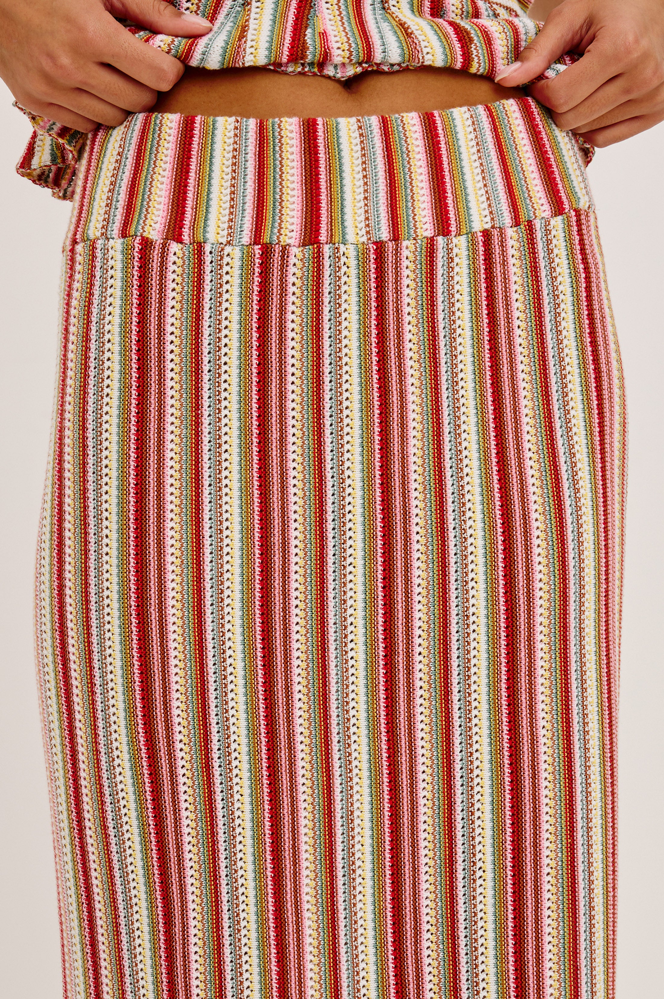 A person wearing the Rails Nari Skirt—a high-waisted maxi with red, yellow, green, and white cotton knit stripes—paired with a matching cropped top. Only their midsection and hands are visible.