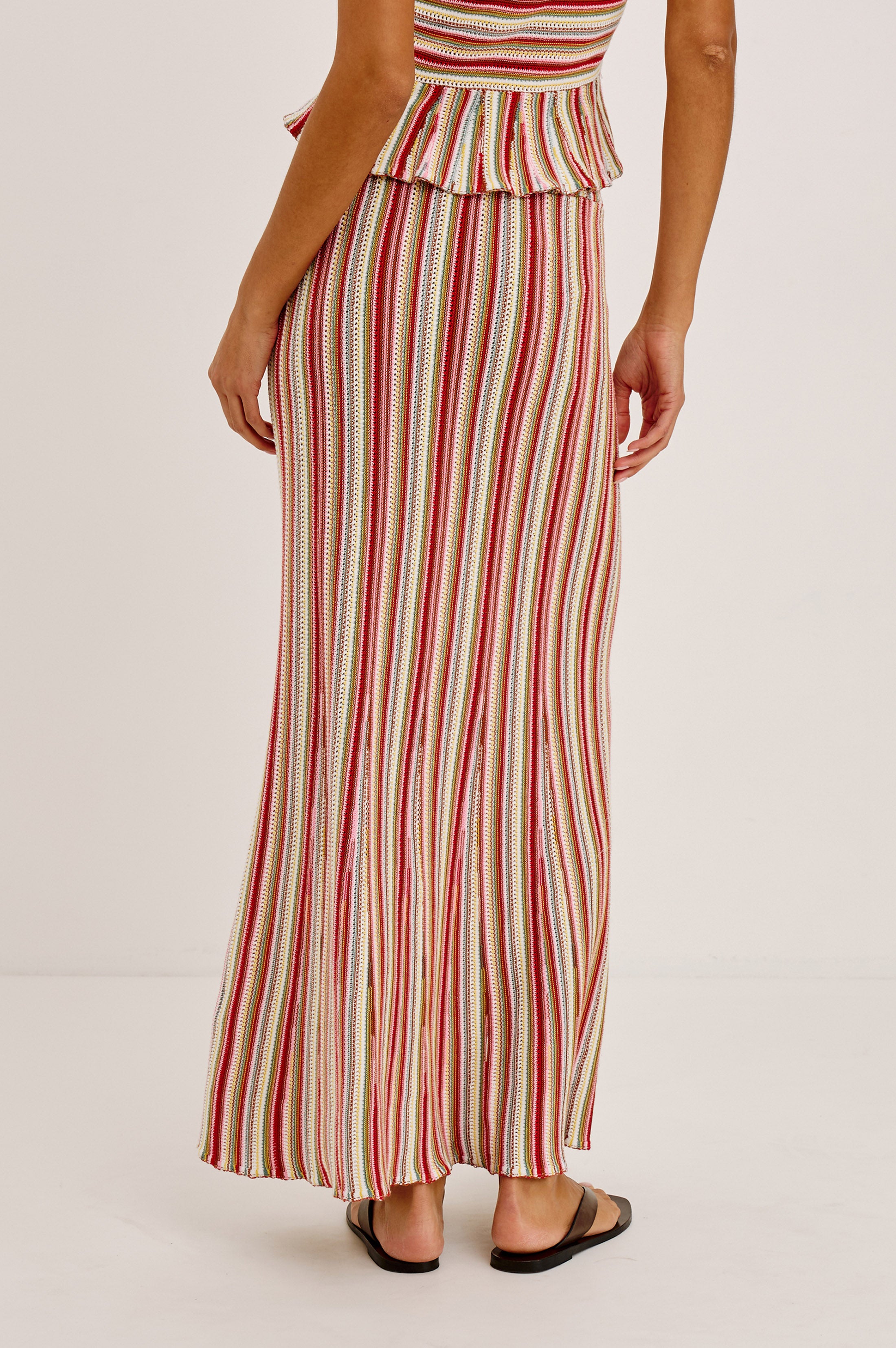 A person faces away wearing the Rails Nari Skirt, a high-waisted maxi with red, white, and green vertical stripes and a ruffled top edge, paired with black flat sandals.